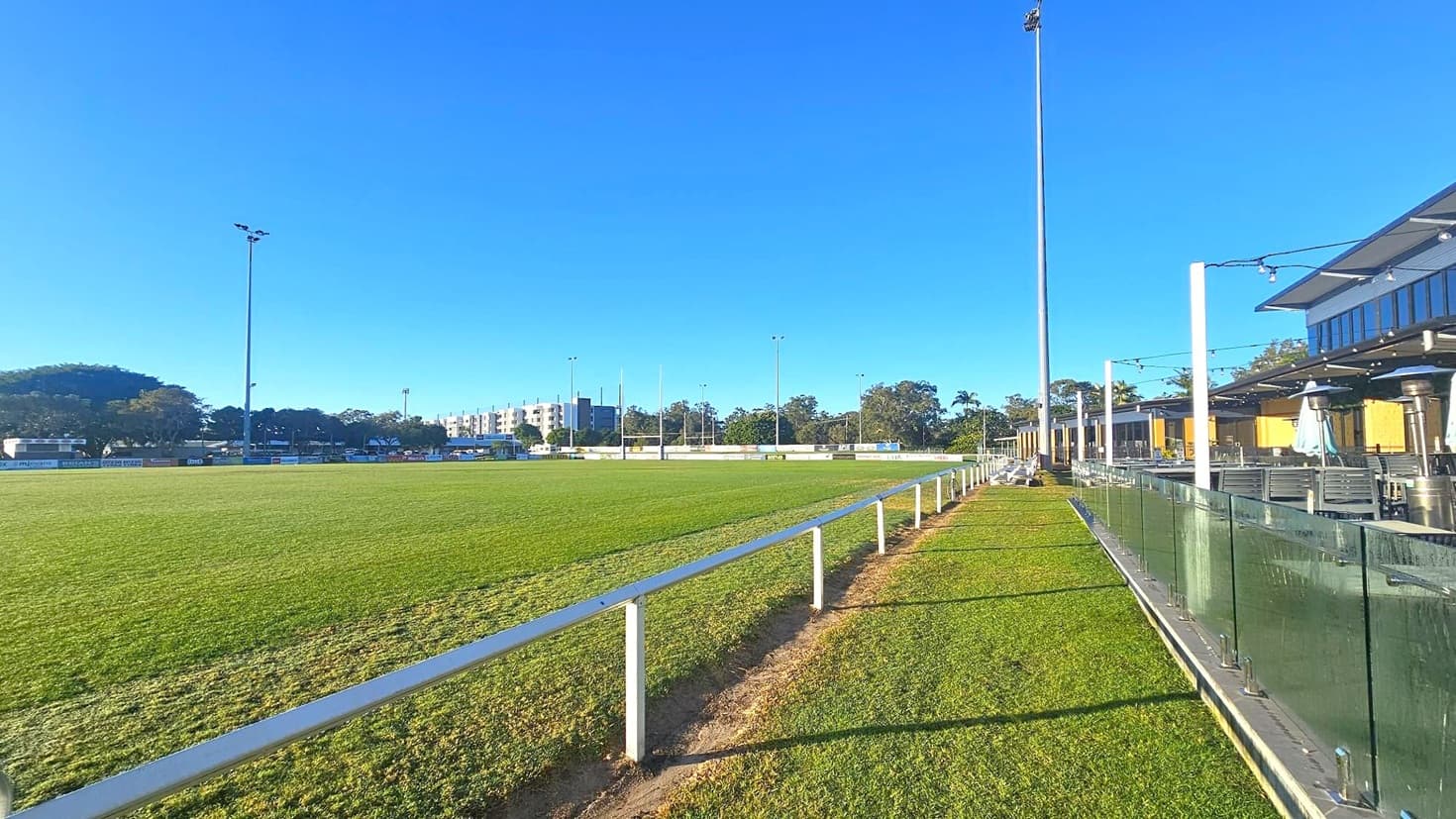 A grassy sports field stretches across the image, bordered by a white railing on the right. Tall floodlights are positioned around the field. On the right, a building with a glass barrier and outdoor seating is visible. In the background, there are trees and a few buildings, including a multi-storey structure. The sky is clear and blue.