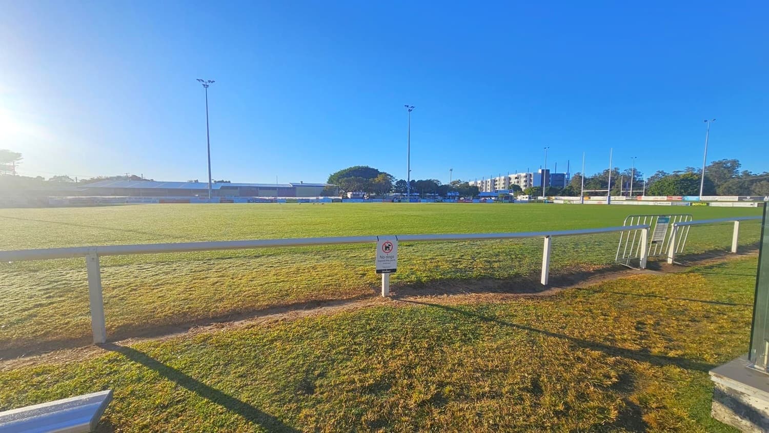 A grassy sports field with a white railing in the foreground. A sign on the railing indicates a restriction. Tall floodlights are positioned around the field. In the background, there are trees, a few buildings, and a row of goalposts. The sky is clear and blue, and the sun is low, casting long shadows across the grass. A few barriers are placed near the railing on the right side.