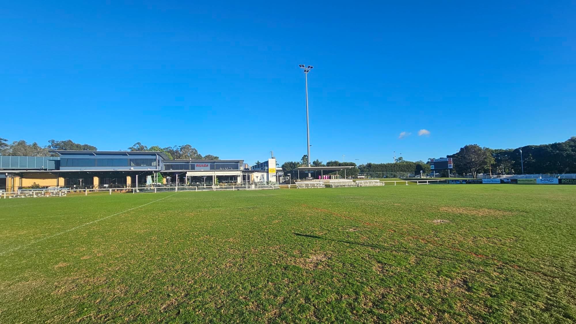 A grassy sports field with a large building in the background on the left, featuring a modern design with a flat roof and large windows. To the right, a tall floodlight pole stands prominently. The field is bordered by a white fence, and there are trees and a scoreboard visible in the distance. The grass appears slightly patchy in areas.