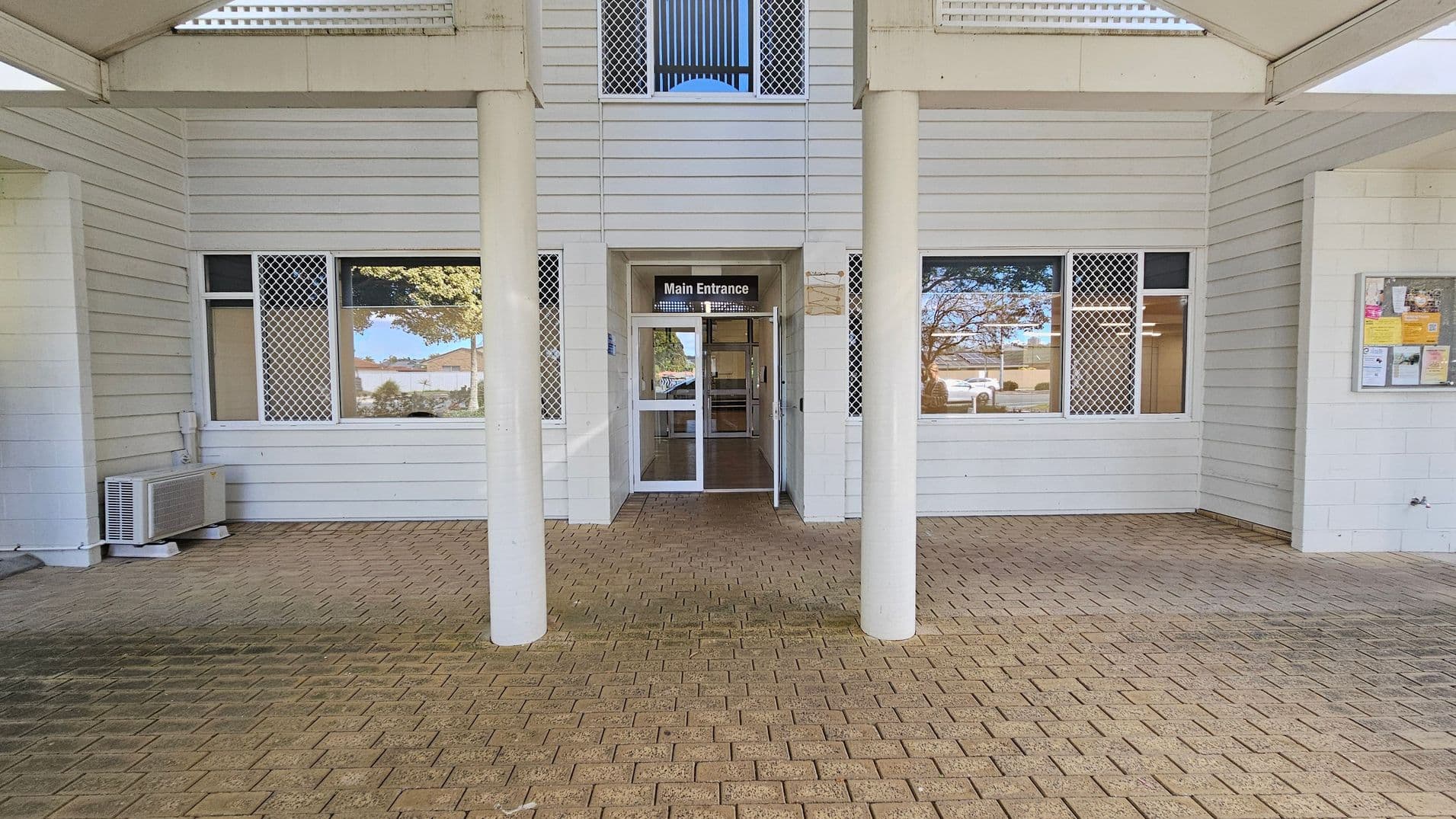 A building entrance with a sign reading "Main Entrance" above glass double doors. The exterior features white horizontal siding and two large windows with security grilles on either side of the doors. An air conditioning unit is visible on the left. The floor is paved with light brown bricks. A notice board with various papers is mounted on the right wall. The ceiling is covered with a light-coloured material, and there are two white columns in front of the entrance.