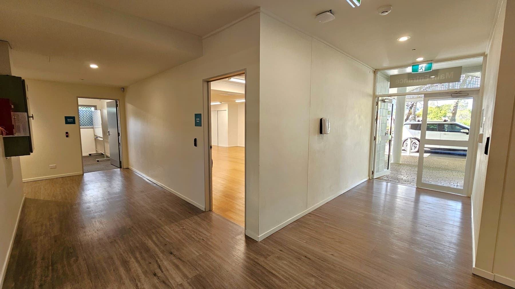 A hallway with wooden flooring and cream-coloured walls. On the left, a door leads to a room with a window and a partially visible sink. In the centre, another door opens to a room with light wooden flooring. On the right, a glass entrance door shows a white car parked outside. The ceiling has recessed lighting and a smoke detector. An exit sign is above the entrance door.
