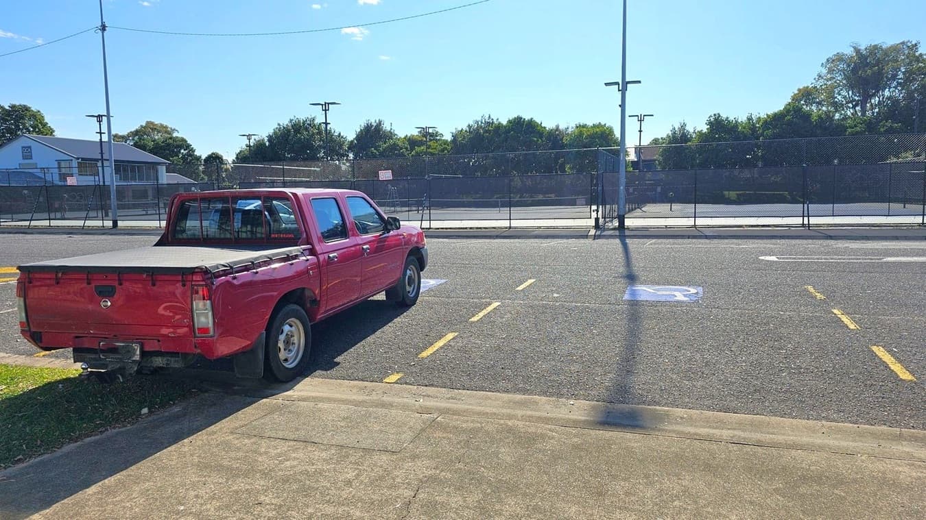 A red pickup truck is parked on the left side of a paved area, partially on the grass. The vehicle is facing away from the camera. In the background, there is a fenced tennis court with trees and a building visible beyond it. The road has a marked disabled parking space and yellow lines. A tall streetlight pole casts a shadow on the ground. The pavement in the foreground is concrete.