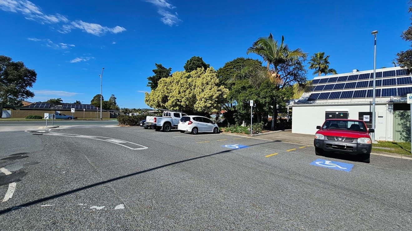 A car park with several vehicles, including a red car in a disabled parking space on the right. A white building with solar panels on the roof is adjacent to the car. Trees with dense foliage are in the background, and a white and a silver vehicle are parked nearby. A road with a car driving past is visible on the left, bordered by a low wall with solar panels. The ground is paved, and a streetlamp is present near the building.