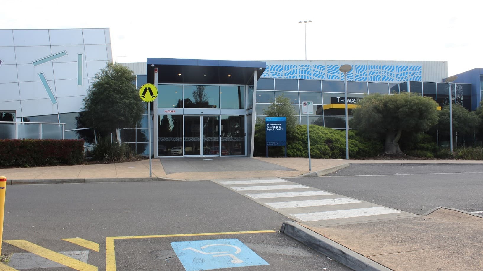 The Thomastown Recreation & Aquatic Centre building is a low-set glass and steel structure with blue and grey accents. On the right, there is a covered waiting area with a grey wooden chair close to a rubbish bin. In front of the facility, there are tall light poles and a sign on the facade that states "Thomastown Recreation Aquatic Center" in white on a black background with white trim. Grass and trees surround the building.
