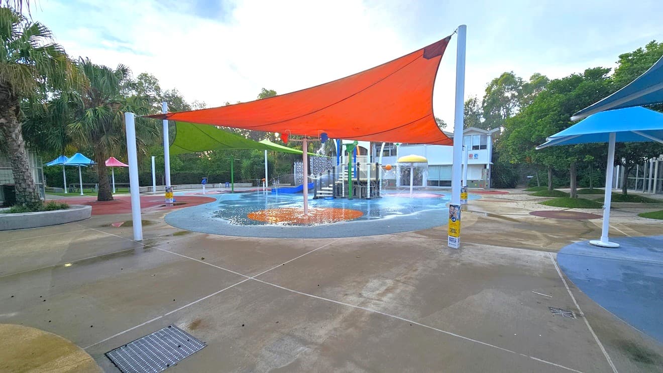 Splash park at Auburn Ruth Everuss Aquatic Centre. In the centre a blue floor with red circular sections for the blue slide with stairs, tipping buckets, water sprays, and a yellow mushroom spray fountain. In front are green and red triangular sun shades wrapped around white metal posts. On the right side is a dedicated seating area with white and blue plastic chairs and blue outdoor umbrella shades. At the back and on the far right side are white buildings with ‘CUMBERLAND COUNCIL’ on its wall.