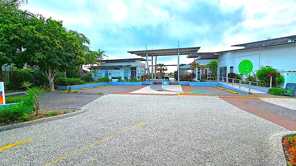 A paved area leads to a building complex with a modern design. On the left, there are trees and shrubs lining the path. In the centre, the entrance features a covered walkway with metal supports. On the right, a white building displays a large green circular logo. The foreground shows a textured pavement with yellow markings, and there are small plants along the edges.