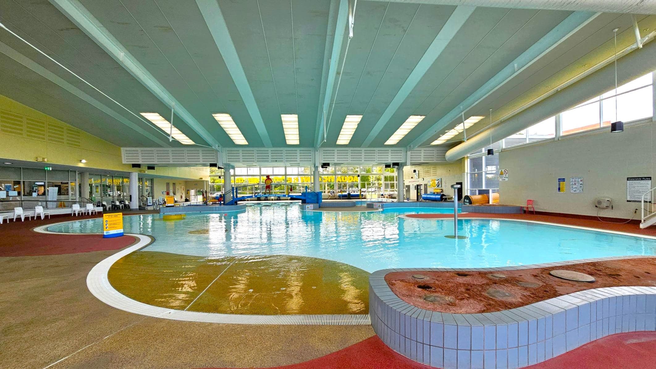 Indoor swimming pool with a curved design. On the left, a lifeguard stands on a blue platform near a small slide. Large windows with yellow signage allow natural light in. The right side features a water slide and a shallow area with a yellow sign. The ceiling has a series of skylights, and the floor is a mix of wet and dry areas with safety markings.
