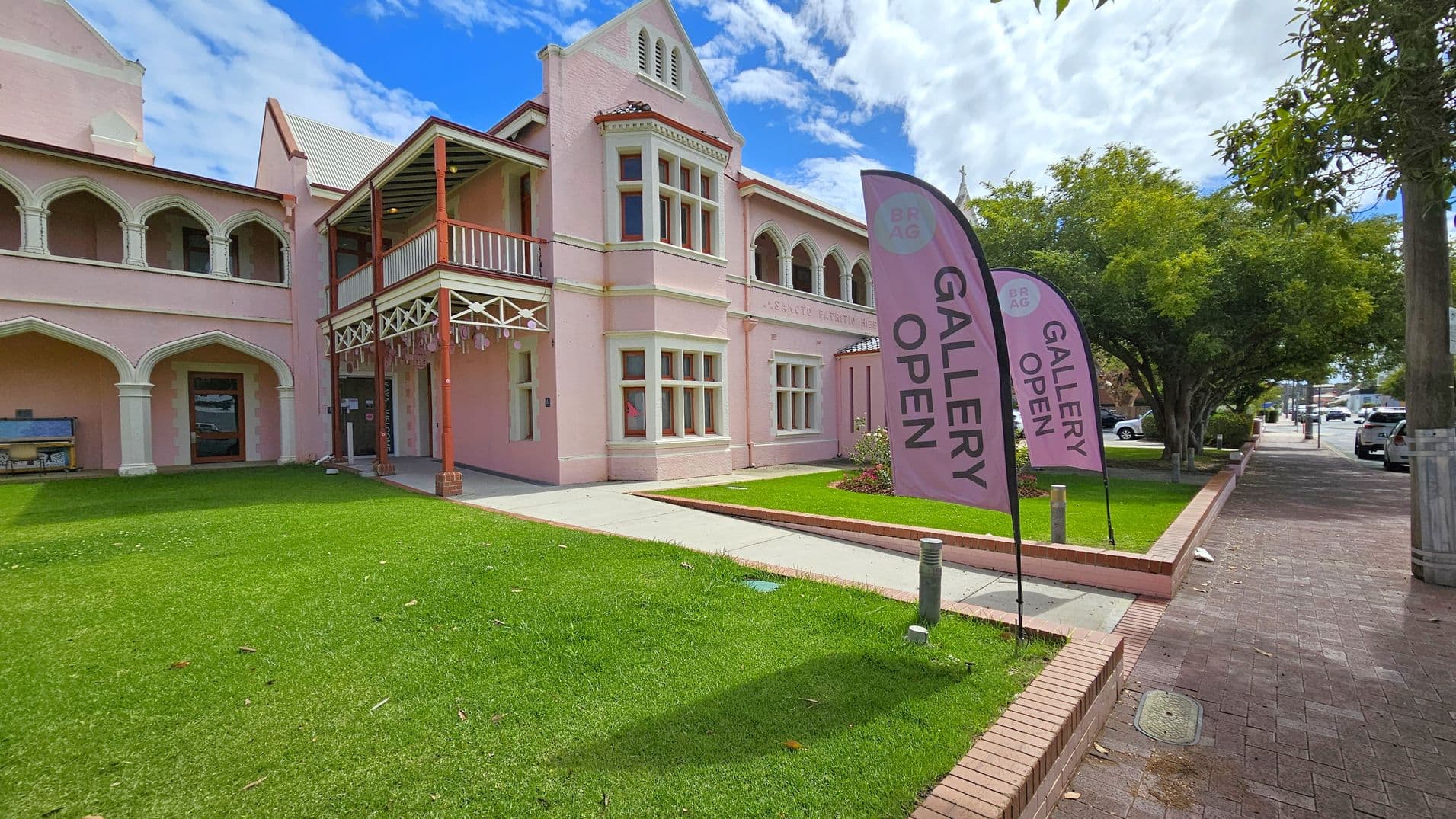 A pink building with arched windows and a cross on top is on the right, with a sign reading "GALLERY OPEN" on a flag. A paved path leads to the entrance, bordered by a green lawn and low brick walls. On the left, a tree overhangs a brick pavement. In the background, there are signs for "ABC" and other buildings, with a few cars visible.