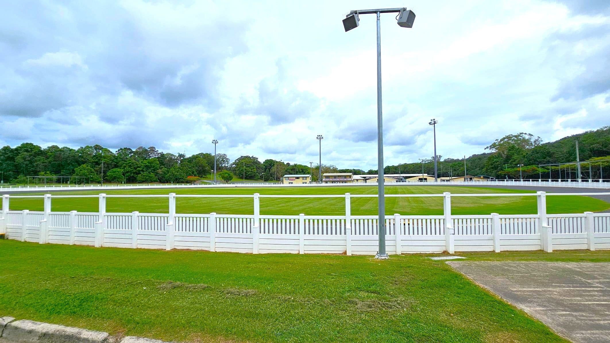A grassy sports field is surrounded by a white picket fence. In the foreground, there is a patch of grass and a concrete path. A tall floodlight stands near the centre. In the background, there are several buildings and more floodlights. Dense trees line the horizon under a cloudy sky.