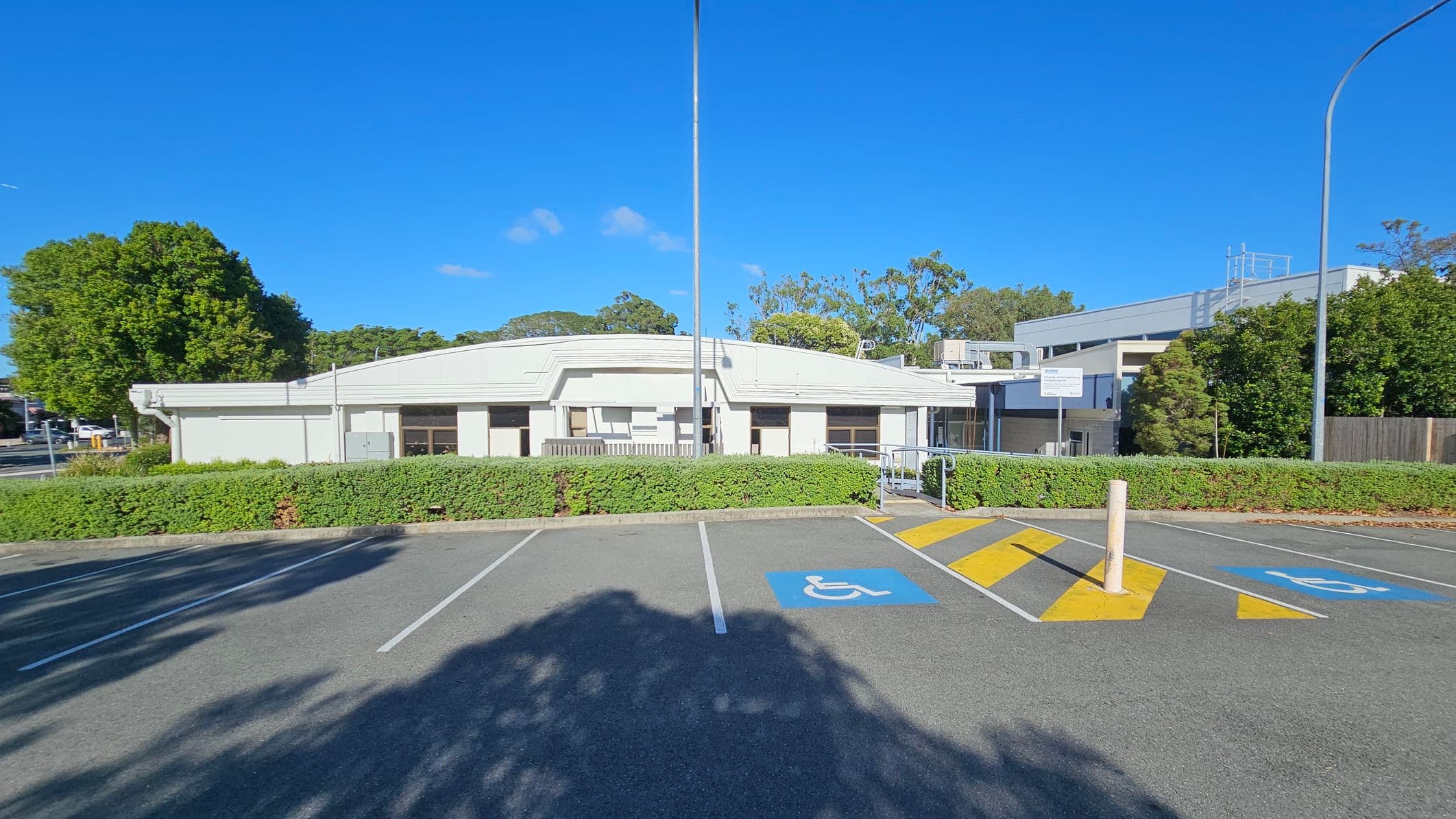 A single-storey white building with a flat roof is in the centre, surrounded by greenery. In the foreground, there is a car park with marked disabled parking spaces and a yellow-striped pedestrian crossing. Tall streetlights are visible on either side. Trees and bushes are in the background, with a clear blue sky above.