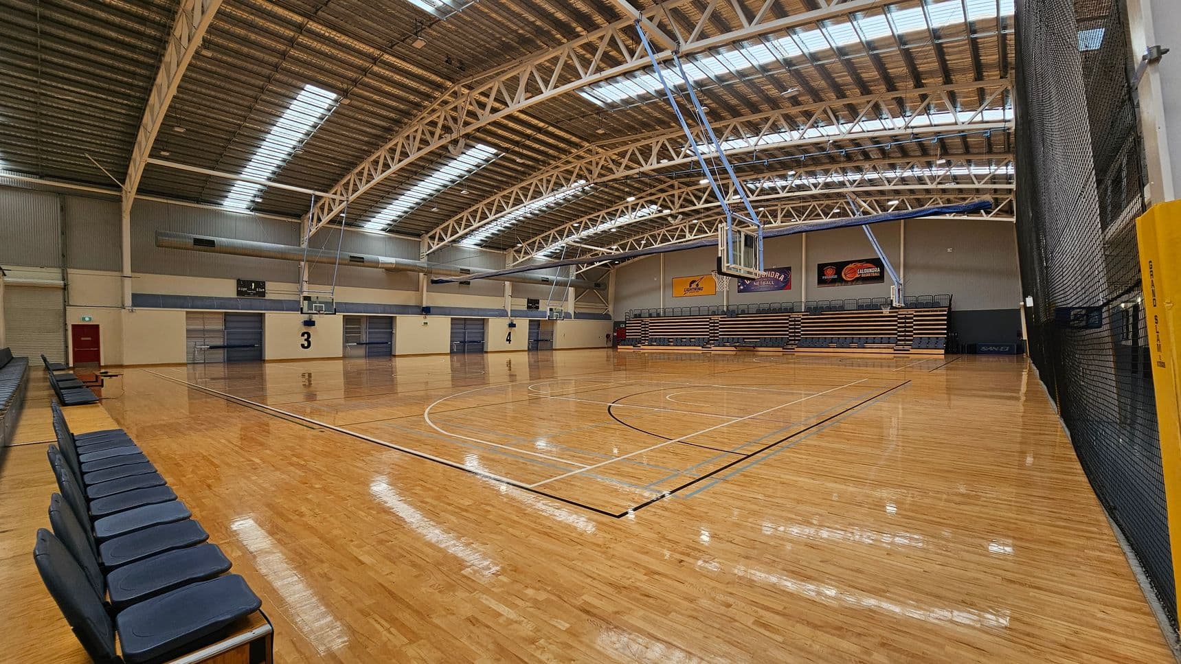 A large indoor sports hall with a polished wooden floor. In the centre, there are two rows of blue chairs facing opposite directions, with "WELCOME" signs at the front. The ceiling features exposed metal beams and skylights. The background shows four basketball courts, each marked with numbers 1 to 4, and basketball hoops. The walls are light-coloured with some banners and doors visible.