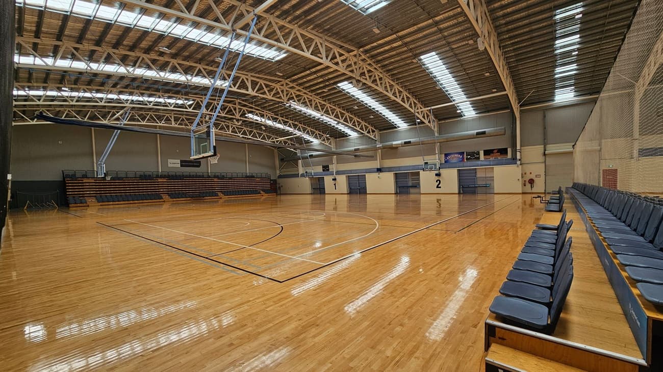 A spacious indoor basketball court with a polished wooden floor. On the left, there are retractable bleachers with dark seating. The right side features more seating along the wall. The ceiling is high with exposed metal beams and skylights, allowing natural light to illuminate the space. Two basketball hoops are visible, one on each end of the court. The walls are light-coloured with some signage and markings indicating court numbers.