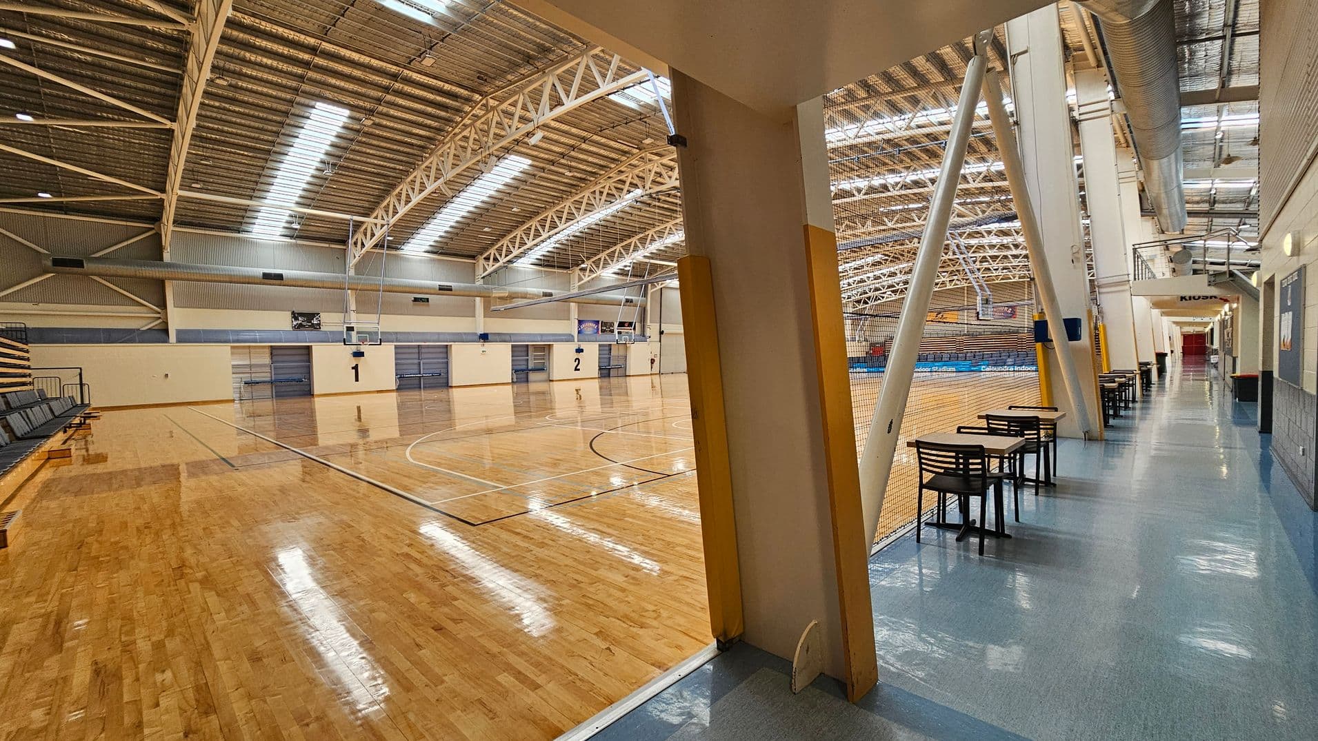 A large indoor sports hall with a polished wooden floor on the left, featuring two basketball courts marked with numbers 1 and 2. The ceiling is high with metal beams and skylights. On the right, a corridor with a blue floor has tables and chairs lined up against a netted barrier. The corridor leads to a distant area marked "Kiosk." The walls are light-coloured, and there are exposed ducts and structural supports visible.