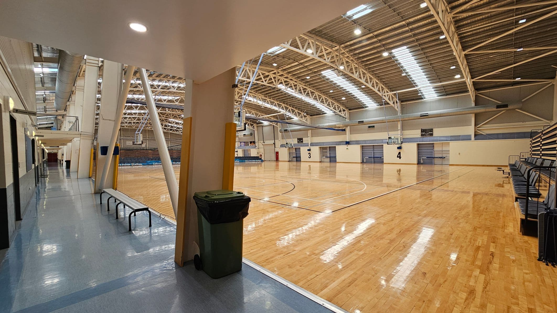 A large indoor sports hall with a polished wooden floor and high, metal truss ceiling. On the right, there are rows of retractable bleachers. In the centre, basketball hoops are visible, and the court is marked with lines. On the left, a corridor with a shiny blue floor leads to the hall, featuring a bench and a rubbish bin. The ceiling has skylights and industrial lighting.