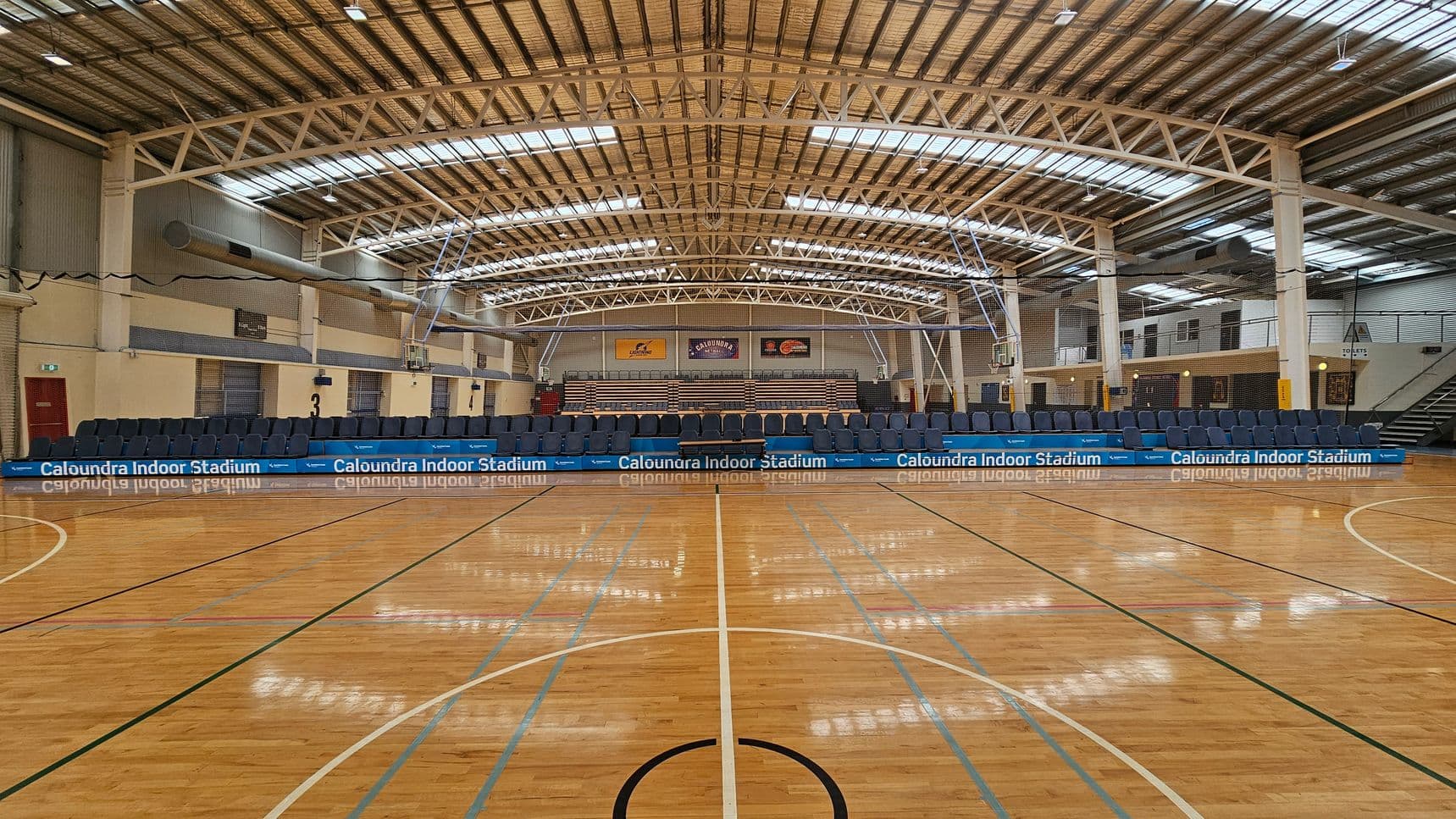 A spacious indoor sports stadium with a polished wooden floor marked for basketball. In the background, there are rows of blue seats and a scoreboard. The ceiling is high with metal beams and skylights, allowing natural light to illuminate the space. Banners are displayed on the far wall above the seating area. The walls are a mix of brick and metal panelling, and there are doors and staircases on the right side.