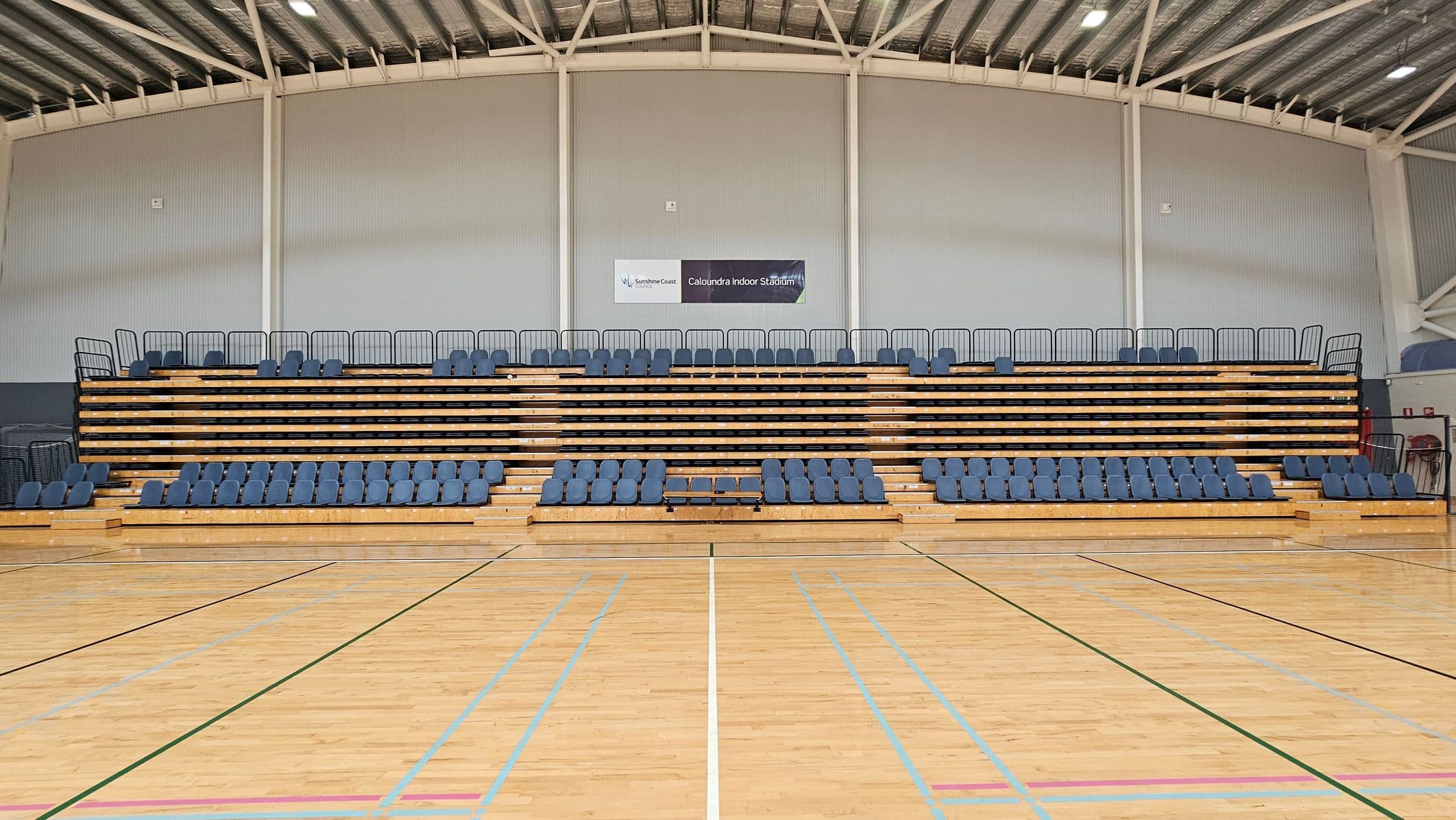 Indoor sports stadium with wooden bleachers featuring blue seats. The bleachers are centred against a grey wall with a sign reading "Caloundra Indoor Stadium" above. The floor is a polished wooden sports court with various coloured lines. The ceiling is high with exposed beams and lighting fixtures.