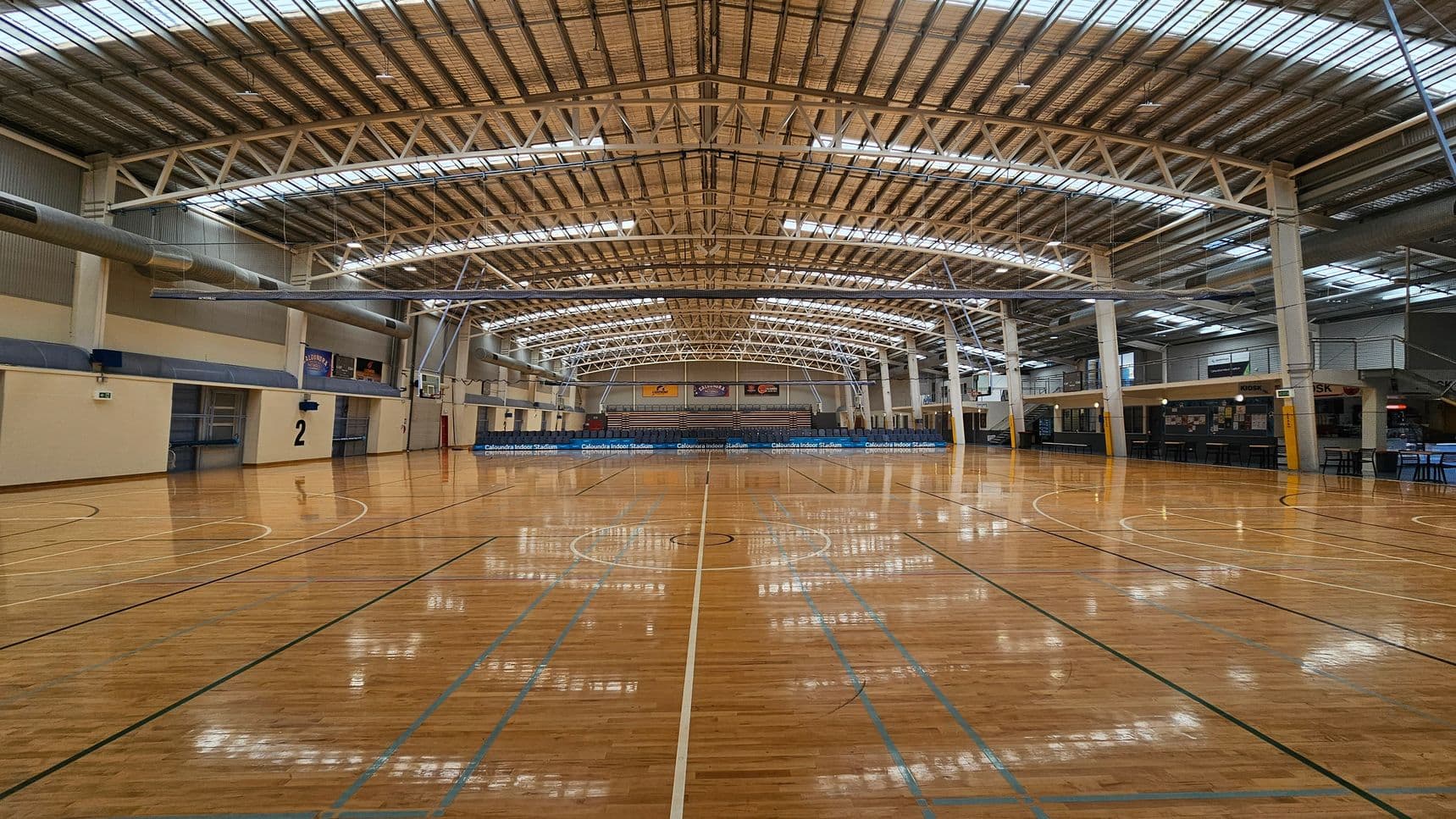 A spacious indoor sports stadium with a polished wooden floor marked for basketball. In the background, there are rows of blue seats and a scoreboard. The ceiling is high with metal beams and skylights, allowing natural light to illuminate the space. Banners are displayed on the far wall above the seating area. The walls are a mix of brick and metal panelling, and there are doors and staircases on the right side.