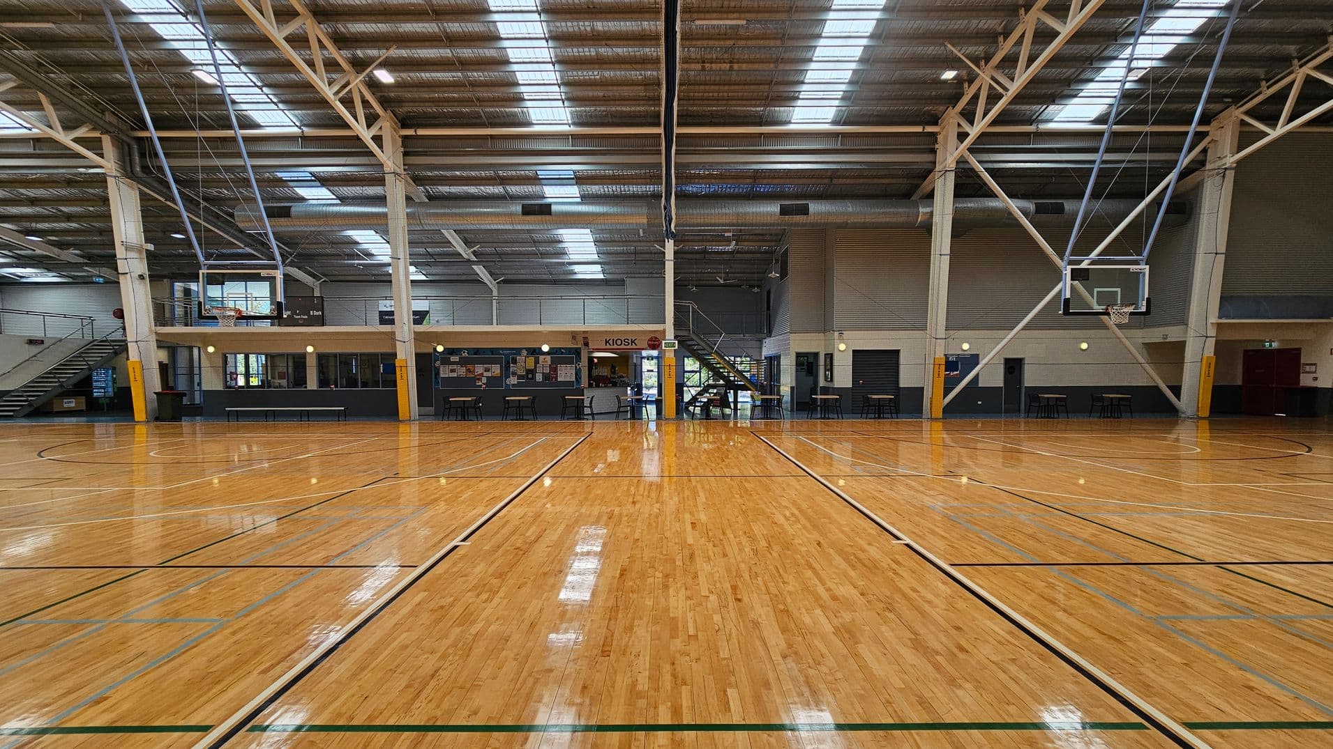 A large indoor sports stadium with a polished wooden floor marked for basketball. The ceiling is high with exposed beams and skylights. On the left, there are numbered walls and a basketball hoop. On the right, there is a kiosk area with tables and chairs. In the background, rows of blue seating are visible, along with banners and signs. The space is well-lit with natural light from the skylights.