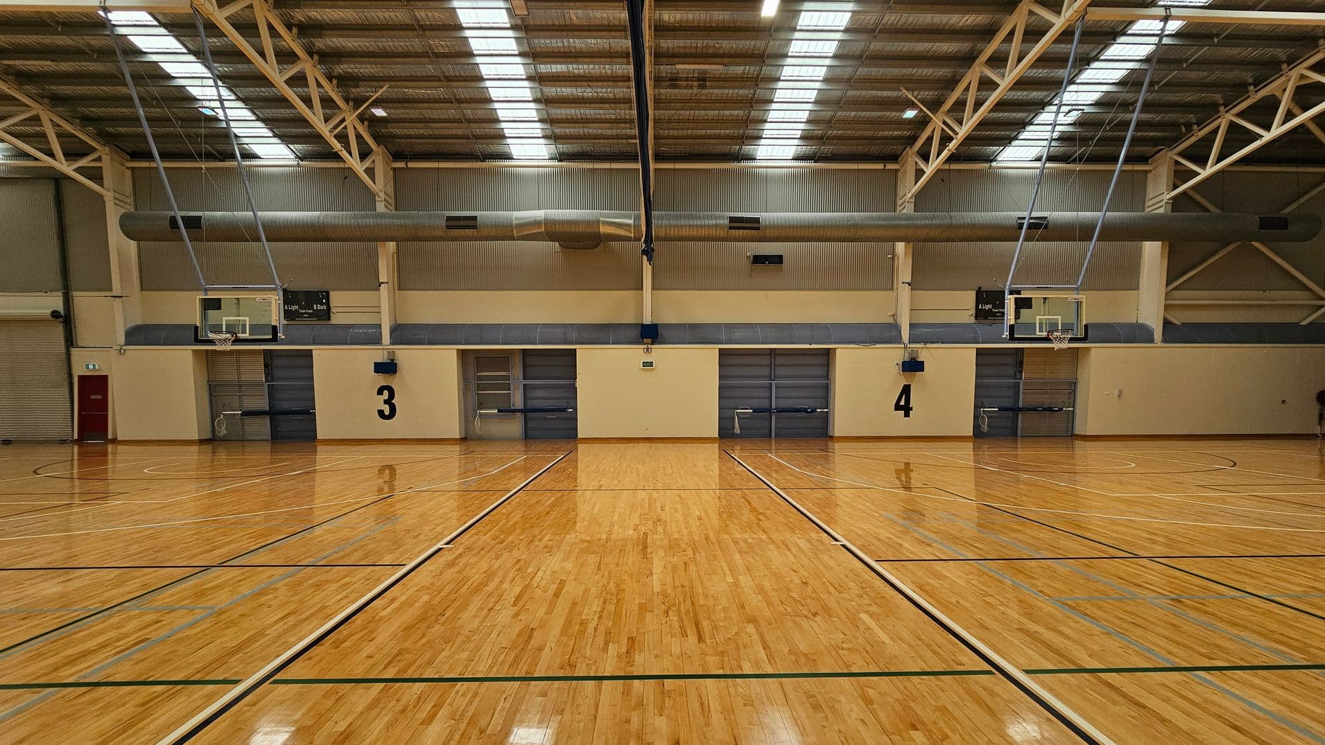 A spacious indoor basketball court with a polished wooden floor marked with lines. Two basketball hoops are visible on either side. The ceiling is high with metal beams and skylights. In the background, there is a kiosk area with tables and chairs, and a staircase leading to an upper level. The walls are a mix of brick and metal panelling.
