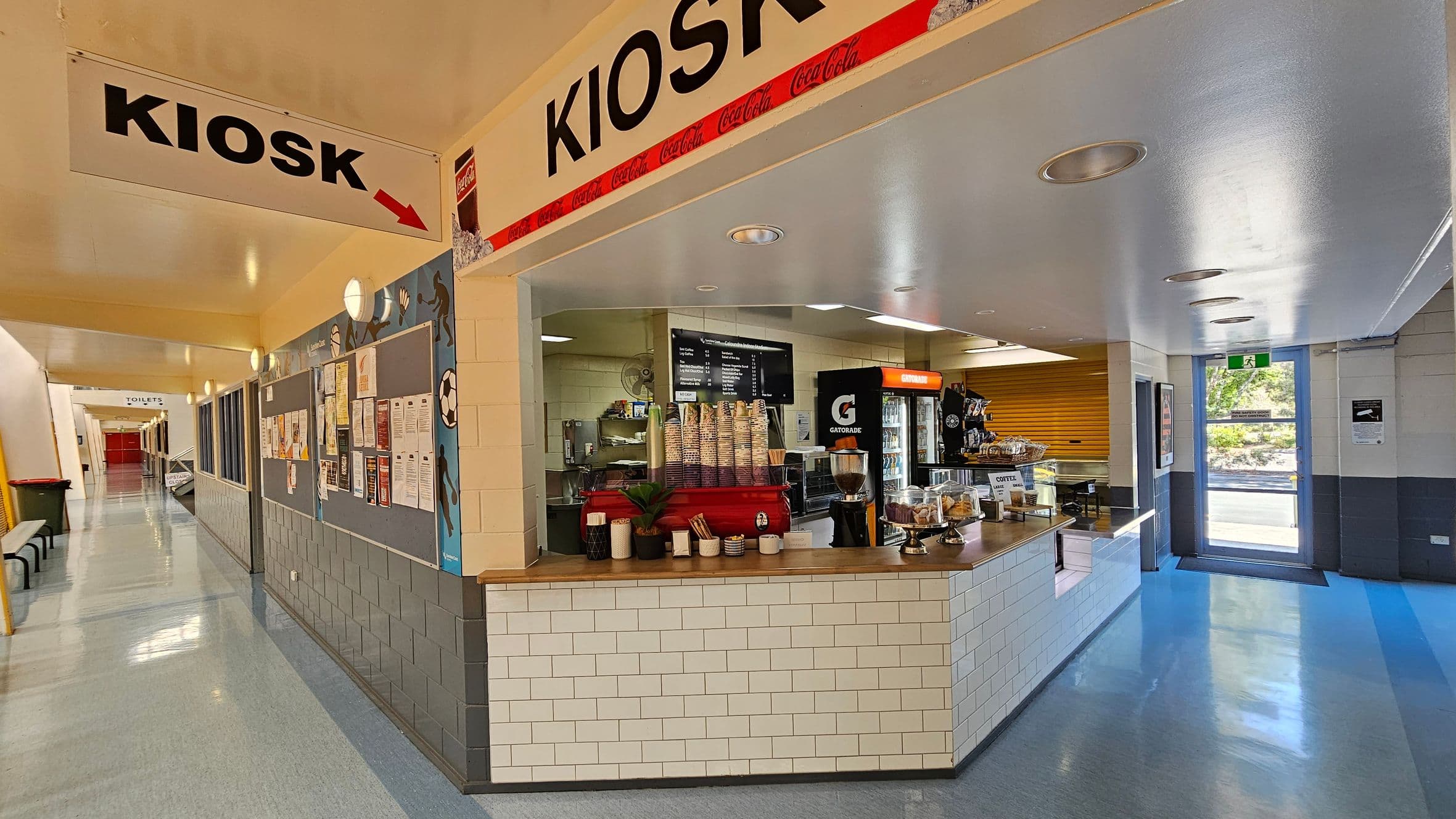 A sports hall with a polished wooden floor and a high ceiling with metal beams and skylights. In the background, there is a kiosk area with tables and chairs. A scoreboard is mounted above, displaying team scores. The wall features notice boards with various posters and information. A yellow pillar stands in the centre, and a staircase is visible on the right leading to an upper level. A green bin is on the left side.