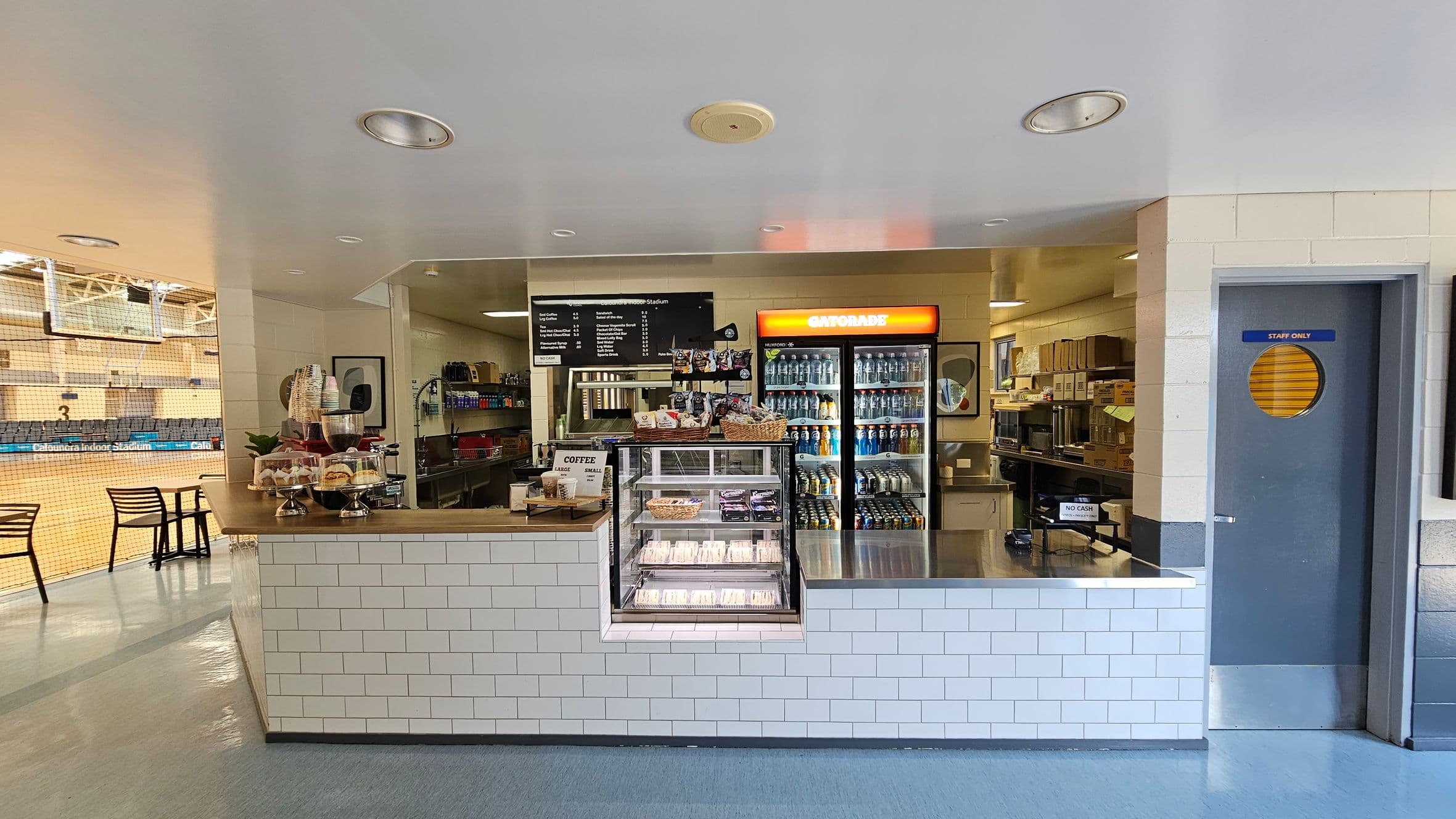 A café counter with a white tiled front. On the left, there are pastries and coffee cups on display. In the centre, a menu board is above a glass case with sandwiches. A fridge with a Gatorade sign is filled with drinks. To the right, a "Staff Only" door is visible. The background shows a sports court with seating and a net. The floor is a mix of grey and blue tiles, and the ceiling is white with recessed lighting.