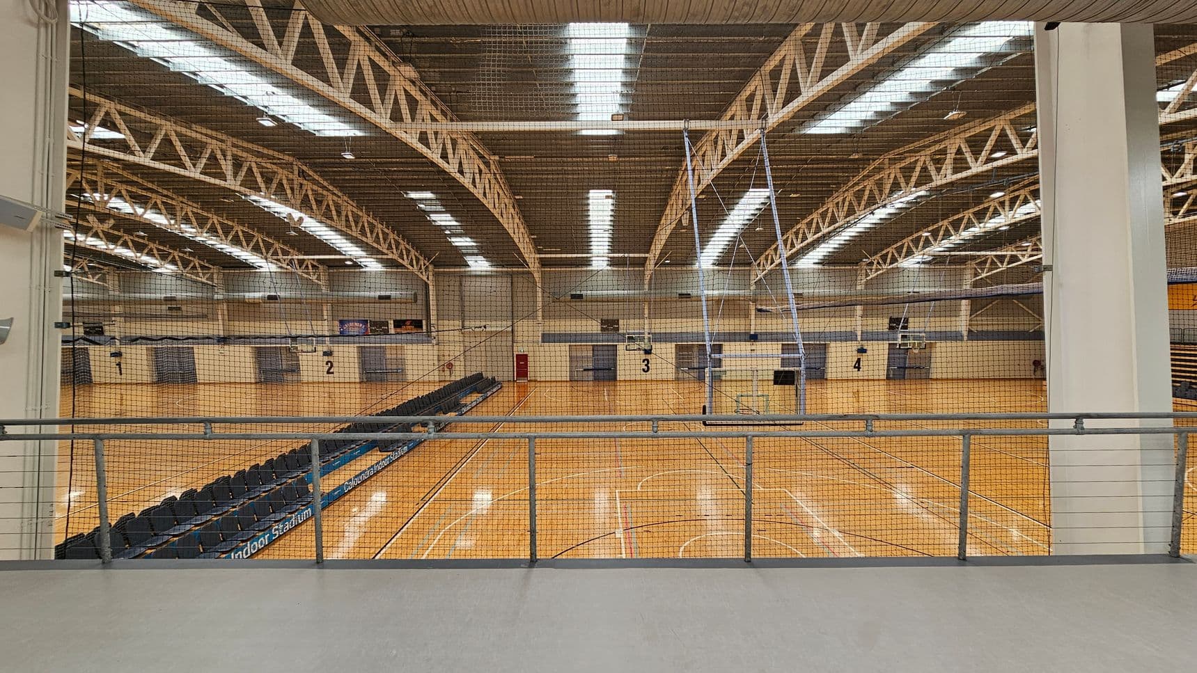 A large indoor sports hall with a wooden floor and multiple basketball courts marked with lines. The ceiling features exposed metal trusses and fluorescent lighting. On the left, there are rows of black seating. A netting barrier runs across the middle, separating the viewing area from the courts. The walls are light-coloured with numbered sections. A metal railing is in the foreground.