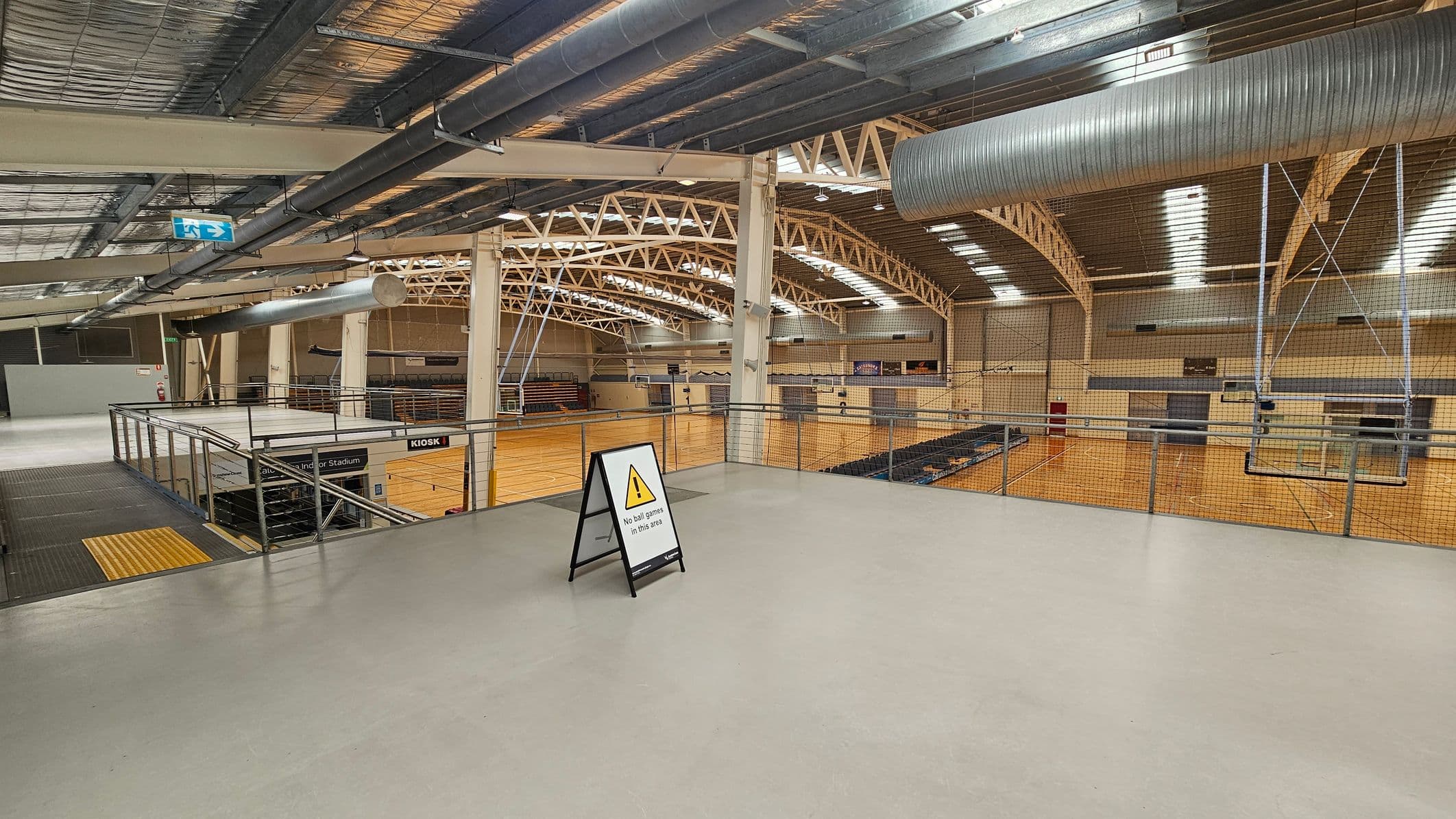 A large indoor sports hall with a high ceiling featuring exposed beams and ductwork. The floor is light grey, and a sign in the foreground reads "No ball games in this area." On the left, there is a staircase leading down, with a "Kiosk" sign visible. The background shows a wooden sports court with seating areas and a netted barrier. An exit sign is visible on the ceiling.