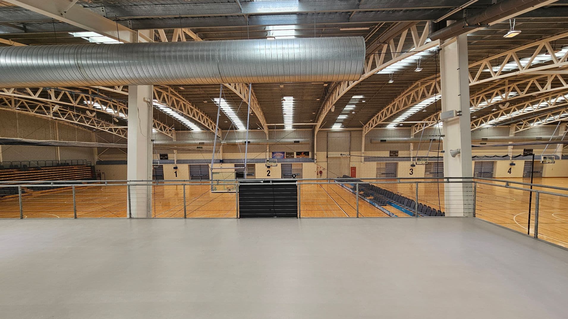 A large indoor sports hall with a high ceiling featuring exposed beams and a prominent metal ventilation duct. The floor is wooden, marked with lines for various sports. On the left, there are tiered seating areas, while on the right, more seating is visible. The hall is divided into multiple courts, numbered 2, 3, and 4, with basketball hoops and nets. A railing is in the foreground, separating a viewing area from the courts below.