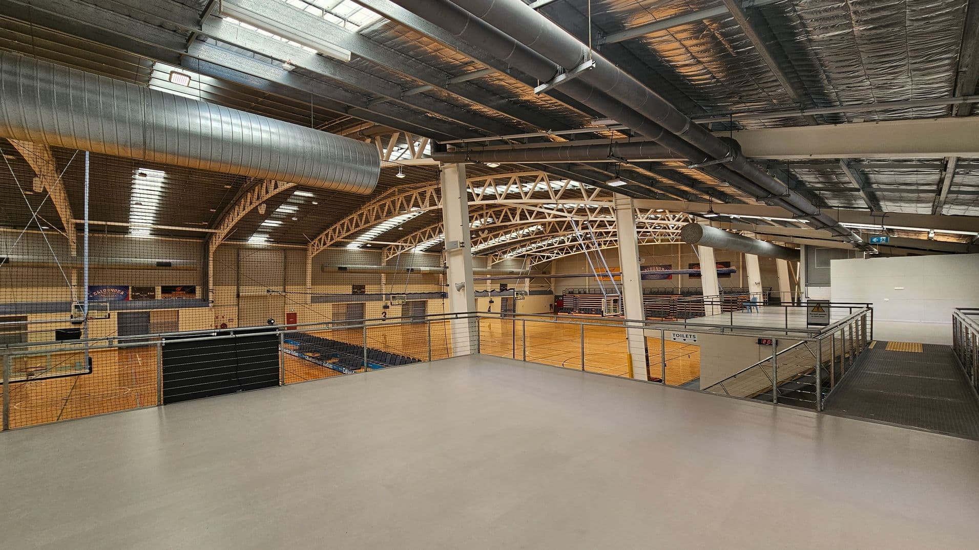 Interior of a large sports hall with a high ceiling featuring exposed metal beams and ductwork. The floor is wooden, marked for sports activities, and surrounded by seating areas. On the left, there are multiple courts with netting above. On the right, a mezzanine level with metal railings overlooks the courts. The ceiling has skylights allowing natural light in. A sign for toilets is visible on the right side near the stairs.