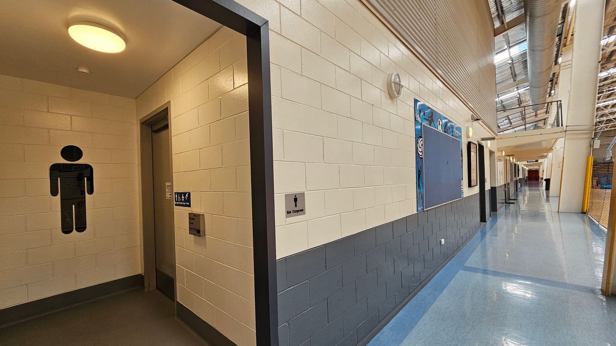 A corridor with a polished floor and a high ceiling. On the left, there is an entrance to a female changing room, marked by a symbol on the wall. Next to it is a closed roller shutter. The walls are tiled in a light colour with a darker base. On the right, a series of doors and a large noticeboard are visible. Ceiling lights illuminate the area, and a sign for "Kiosk 1" is seen further down the corridor. The ceiling has a metal structure with skylights.