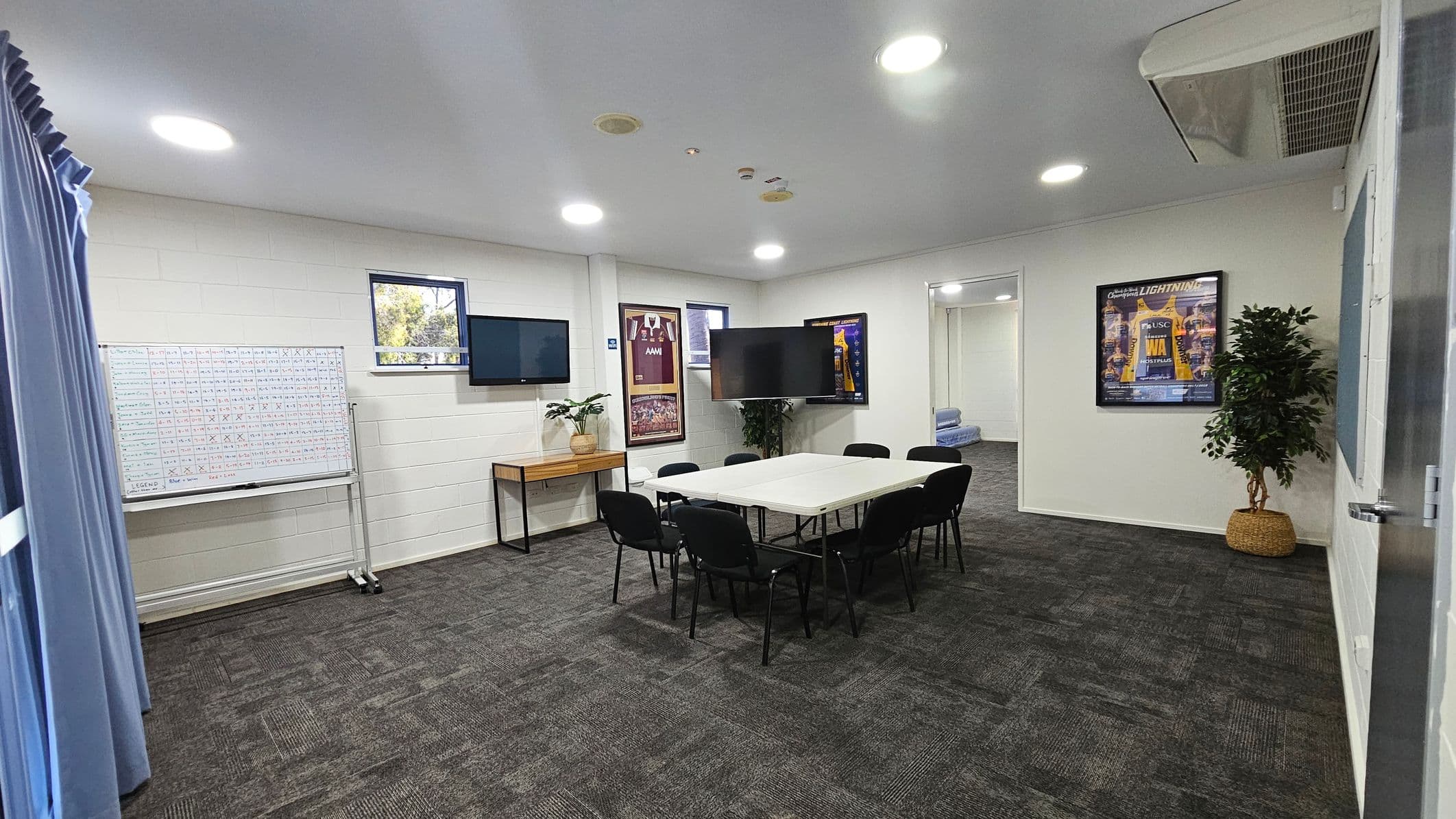 A hallway with a blue and grey tiled floor and cream and grey walls. On the left, an open door leads to a room with a whiteboard and a small window. A framed sports poster is on the wall next to the door. To the right, a closed grey roller shutter is visible. Further down the hallway, there are red double doors and a few black tables and chairs. The ceiling is high with visible beams.