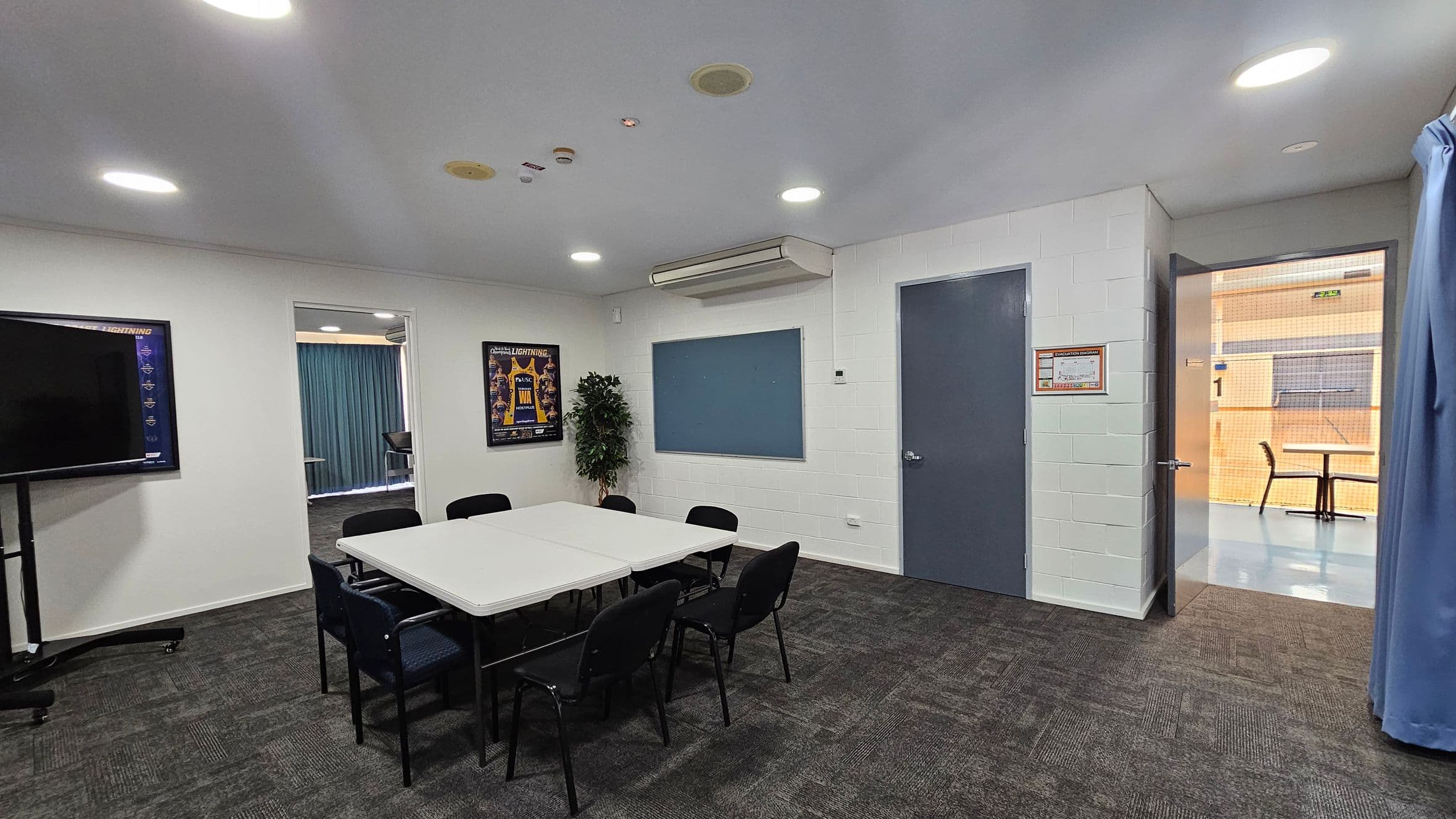A meeting room with a rectangular table surrounded by black chairs on a dark carpeted floor. On the left, a large screen is mounted on a stand. A poster is on the wall next to a doorway leading to another room with blue curtains. In the centre, a blue notice board is mounted on the wall above a potted plant. On the right, a door is open, revealing a hallway with a table and chair. The ceiling has recessed lighting and an air conditioning unit.
