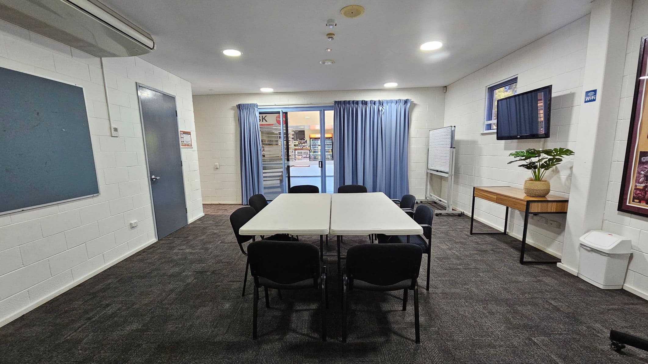 A room with a dark carpeted floor and white walls. In the centre, two rectangular tables are pushed together, surrounded by eight black chairs. On the right, a wooden table holds a potted plant beneath a wall-mounted TV. A whiteboard is nearby. The back wall features blue curtains covering a glass door, leading to a view of a shop. On the left, a grey door and a blue notice board are visible. The ceiling has recessed lighting and an air conditioning unit.