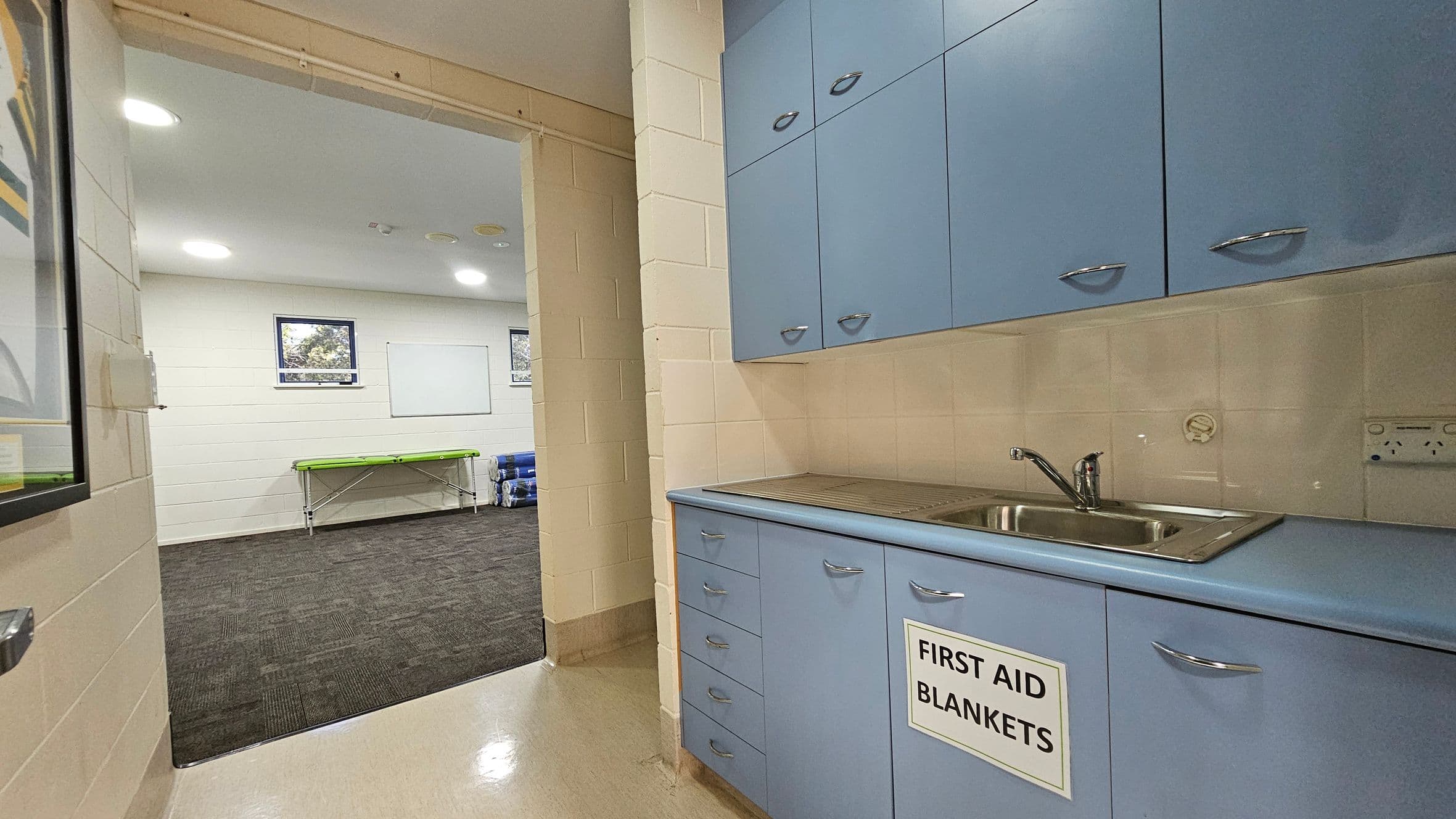 A room with light blue cabinets on the right, featuring a sink and a sign reading "First Aid Blankets." The floor is a light-coloured linoleum. Through an open doorway on the left, a room with dark carpet is visible, containing a green stretcher and rolled-up mats. The walls are cream-coloured with two small windows and a whiteboard. The ceiling has recessed lighting.