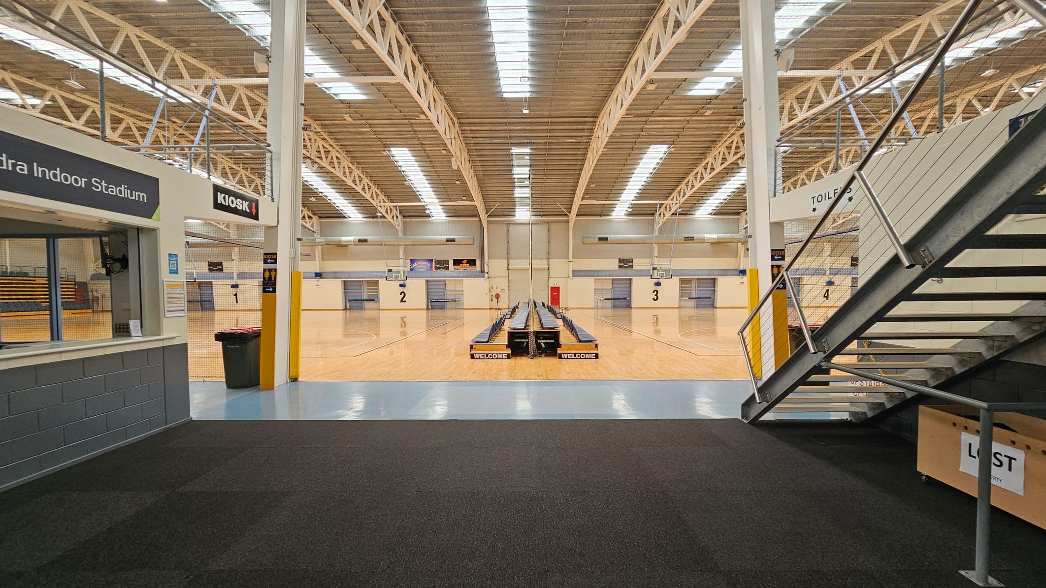 Glass entrance with blue framing, leading into a building. On the left, a yellow poster with social media information. On the right, a sign with entry conditions. Inside, a reception desk is visible, with a staircase to the right. The floor is tiled, and the ceiling has recessed lighting. Outside, a concrete path and some greenery are visible.