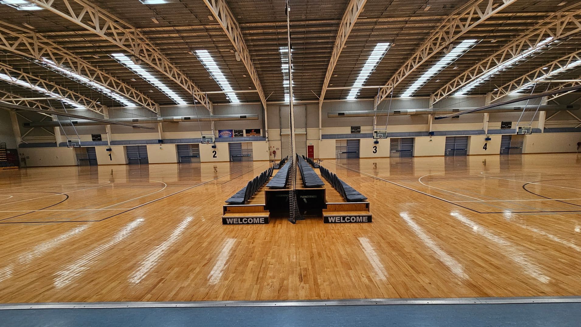 A large indoor sports hall with a polished wooden floor. In the centre, there are two rows of blue chairs facing opposite directions, with "WELCOME" signs at the front. The ceiling features exposed metal beams and skylights. The walls are beige with numbered sections from 1 to 4, each with a basketball hoop. The space is well-lit, and the floor markings indicate multiple basketball courts.