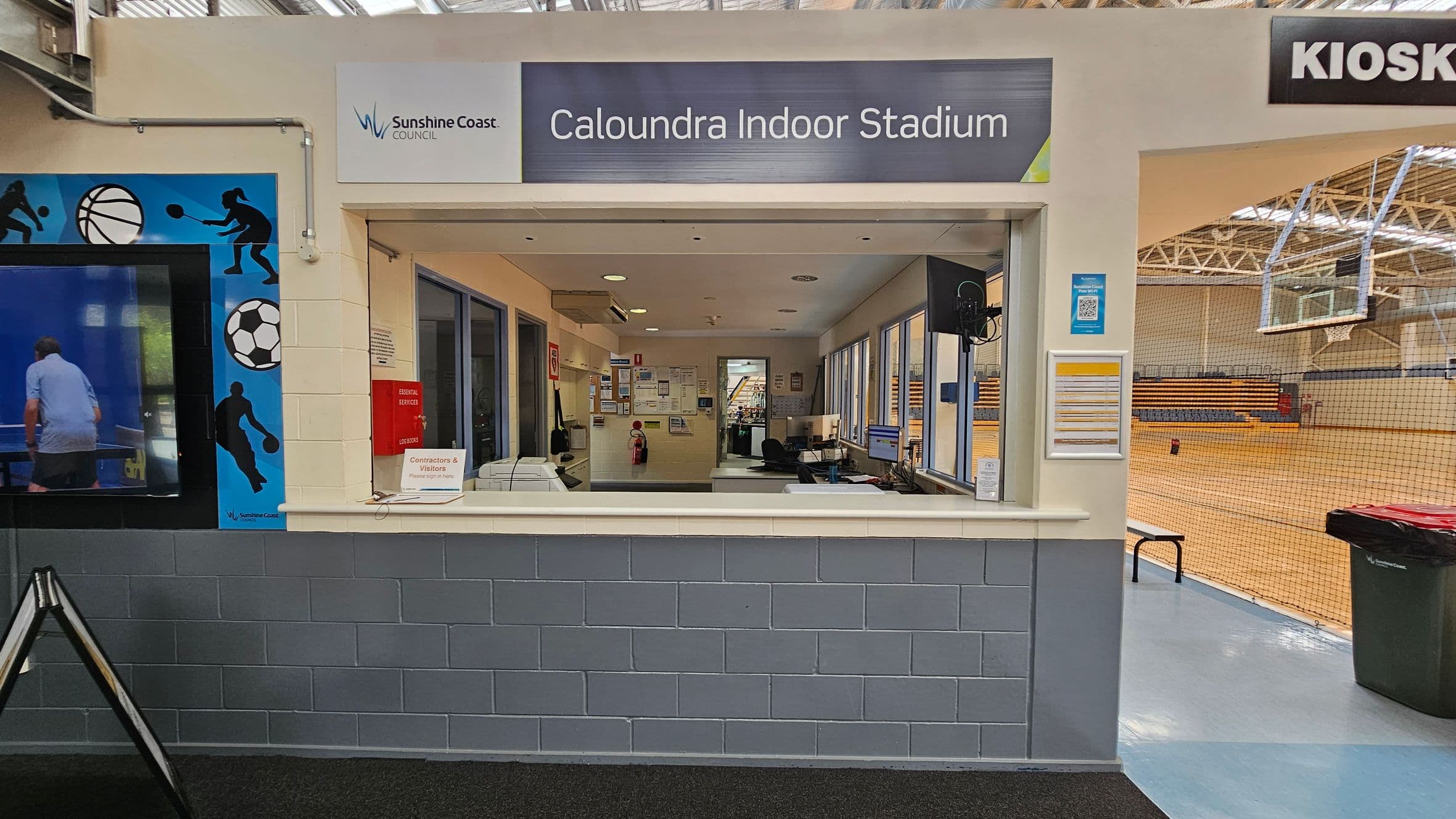 Reception area of Caloundra Indoor Stadium. The counter has a sign for contractors and visitors. Behind the counter are office equipment and notice boards. To the left, a wall features sports-themed graphics. On the right, an entrance leads to a gymnasium with a basketball hoop and bleachers. The ceiling has exposed beams and lights, and the floor is a mix of carpet and polished surface. A kiosk sign is visible above the entrance.