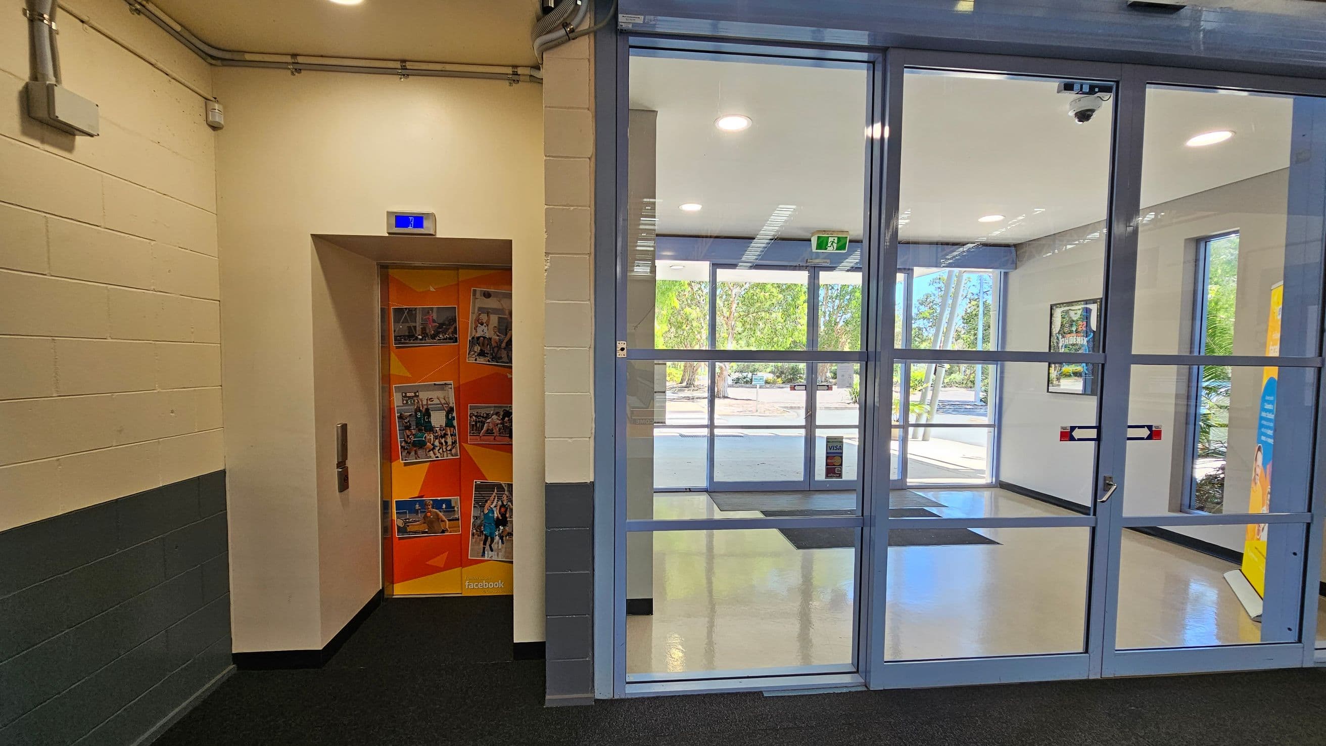 On the left, there's a lift with an orange door featuring various photos. The wall is cream with a grey lower section, and there are visible pipes near the ceiling. On the right, a glass entrance door leads to a bright lobby with large windows showing trees outside. The floor is a mix of dark carpet and light tiles. A poster is partially visible on the far right.