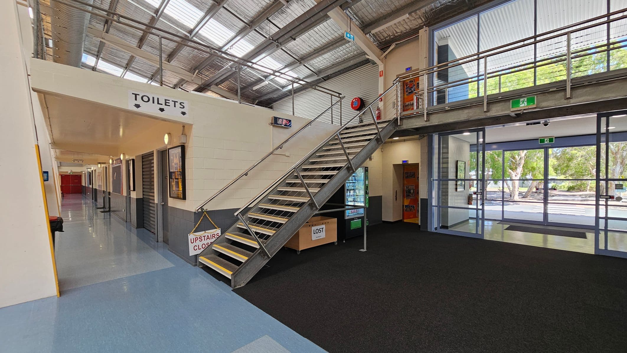 A modern indoor space with a metal staircase in the centre leading to a mezzanine level. To the left, a corridor with a sign for toilets and several doors. The floor is a mix of blue and black tiles. On the right, large glass doors and windows provide a view of trees outside. A vending machine and a "Lost" box are near the staircase. The ceiling is industrial with exposed metal beams and skylights.