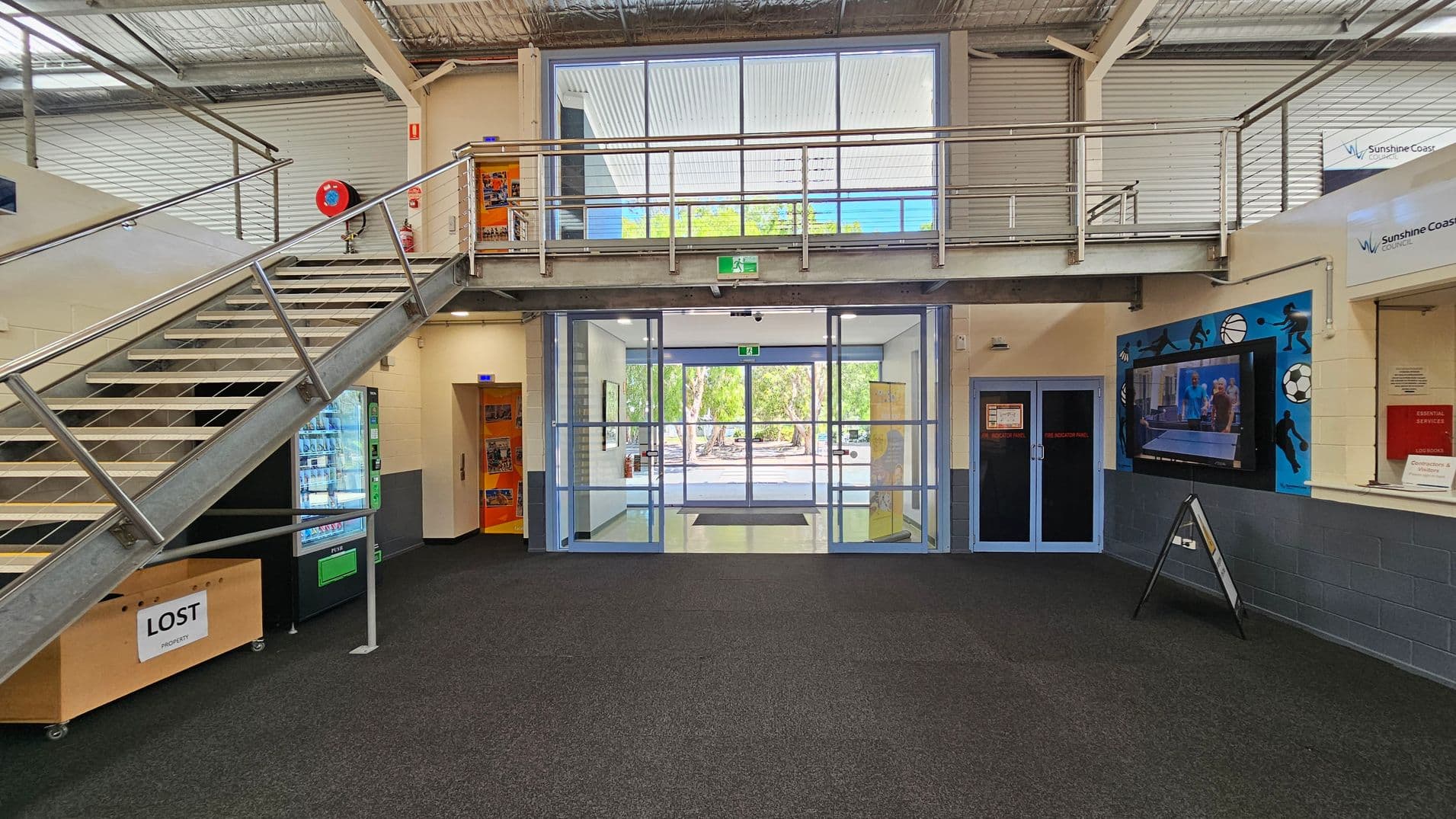 A spacious indoor area with a dark carpeted floor. On the left, a metal staircase leads to an upper level with a railing. Below the stairs, there's a vending machine and a "Lost Property" box. In the centre, large glass doors open to an outdoor area with trees visible. On the right, a wall-mounted TV displays a sports scene, and there's a sign for "Sunshine Coast Council" above. The ceiling is industrial with exposed beams and ductwork.