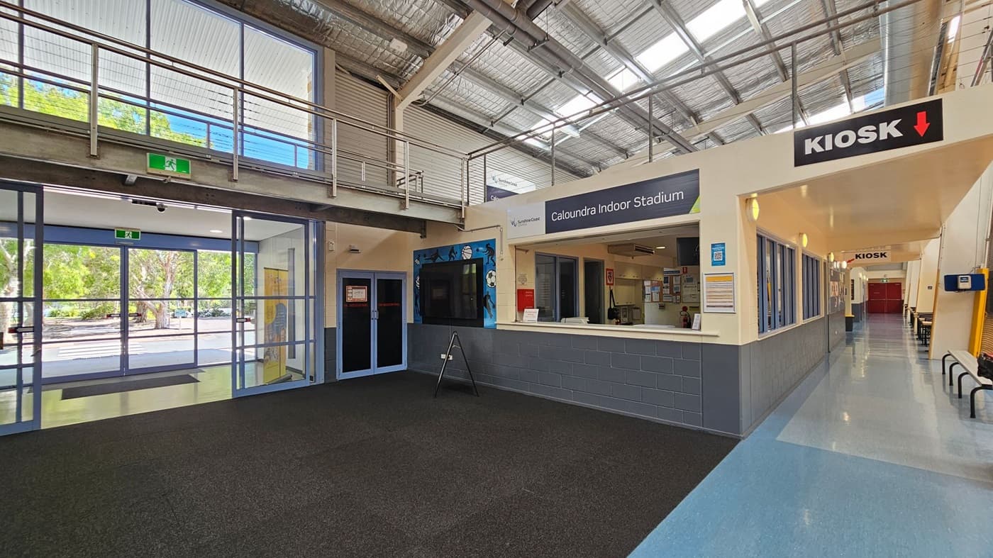 Interior of a building with a high ceiling featuring exposed metal beams and skylights. On the left, large glass doors lead outside, showing trees through the windows. In the centre, a reception area for "Caloundra Indoor Stadium" with a counter and signage. To the right, a corridor with a "Kiosk" sign above, leading to a red door. The floor is a mix of dark carpet and light blue tiles, and there are benches along the corridor.