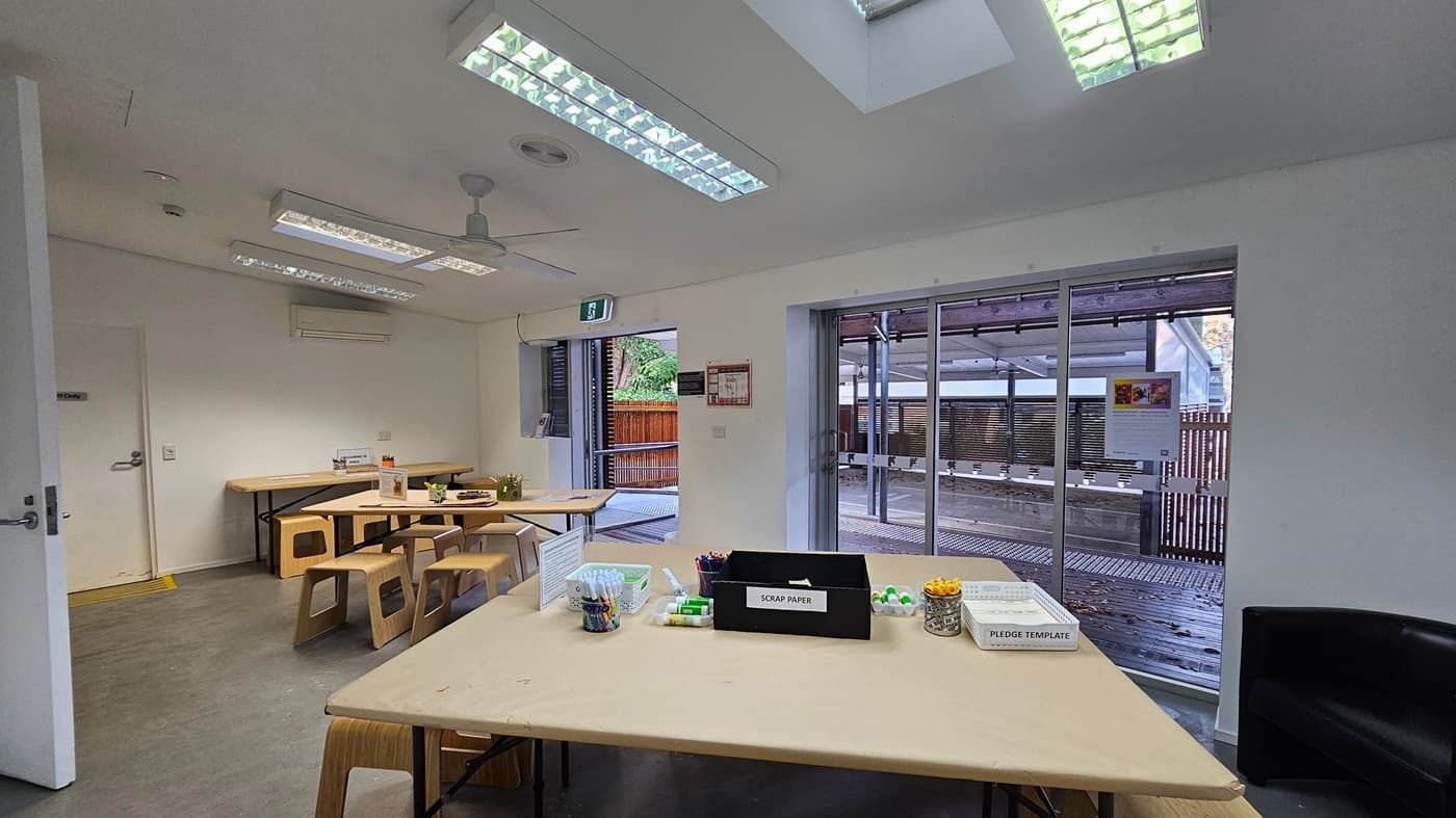 A room with a light grey floor and white walls. In the centre, there are two wooden tables with matching stools. The left side has large glass doors leading to an outdoor area with a wooden fence. A small window is next to the doors. The ceiling features fluorescent lights and a skylight. An exit sign is above the doors. On the right, a partially open door leads to another room. A ceiling fan is also visible.