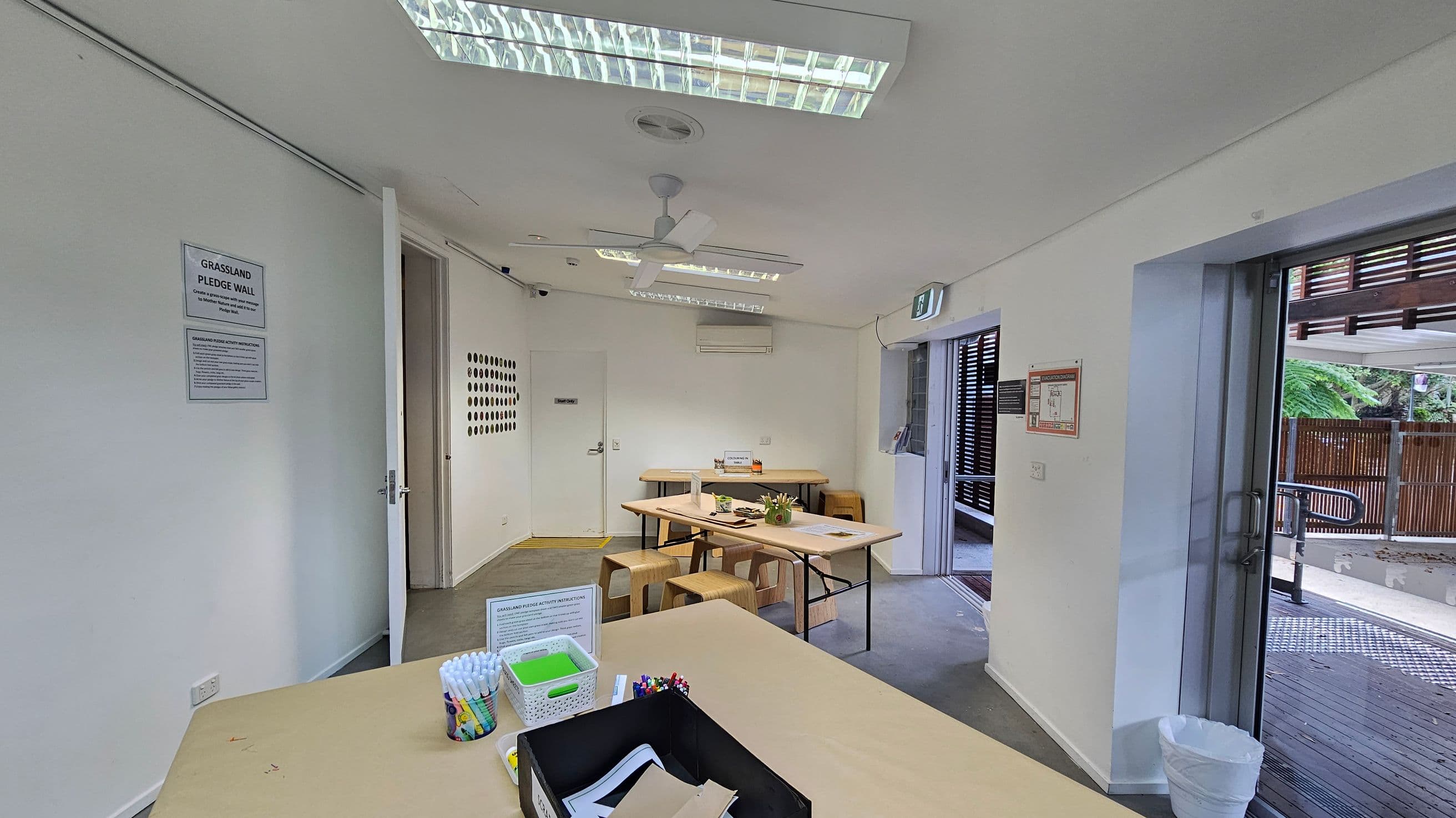 A room with white walls and a ceiling featuring fluorescent lights and a ceiling fan. On the left, a sign reads "Grassland Pledge Wall" with instructions below. In the centre, tables with stools are arranged, holding various craft supplies. A door is open on the left wall, and a window on the right shows an outdoor area with wooden fencing. The floor is grey, and there is a small white bin near the window.