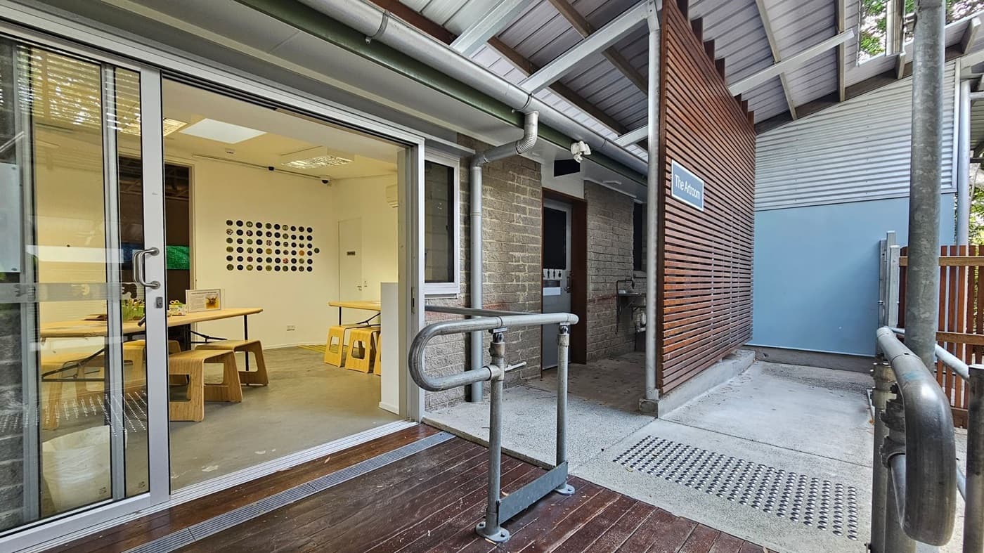 A modern building entrance with a sliding glass door on the left, revealing a room with wooden tables and stools. The floor inside is light grey, and the walls are white with a circular pattern decoration. Outside, there's a wooden deck and a metal railing. On the right, a brick wall and a wooden slatted panel with a sign reading "The Anthoni" are visible. The ceiling is metal with exposed beams, and the exterior wall is partly blue.