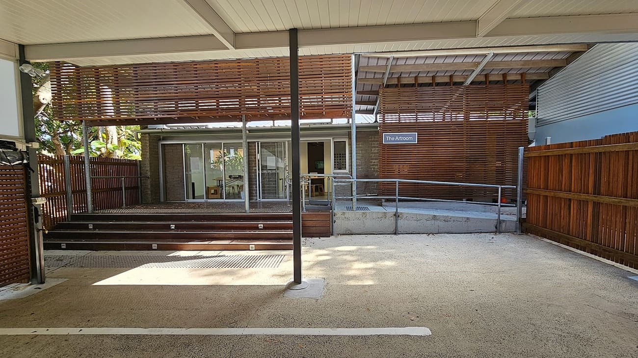 A covered outdoor area with a textured concrete floor and a white ceiling. In the centre, a metal pole supports the structure. Wooden slats form a partial wall on the left and right, with a wooden fence extending on both sides. In the background, a building with sliding glass doors and a sign reading "The Artroom" is visible. Steps lead up to the entrance, and a metal railing runs along the right side. Trees and foliage can be seen through the slats on the left.