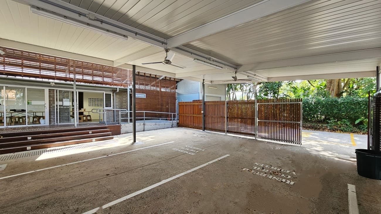 Covered outdoor area with a concrete floor, scattered with dry leaves. The ceiling is lined with panels and has ceiling fans. On the left, there are glass doors and windows leading to an indoor space with tables and chairs. A wooden ramp and steps provide access. On the right, a wooden fence and greenery are visible. The background shows trees and bushes, creating a natural setting.