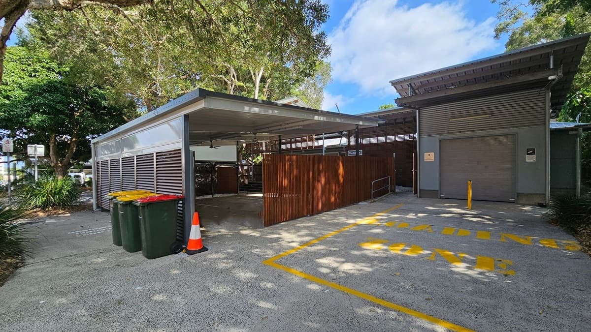 A covered outdoor area with a metal roof and wooden slat walls on the right. Three wheelie bins with yellow and red lids are on the left, next to an orange traffic cone. The ground is concrete with a marked "LOADING ZONE" in yellow. A large roller door is on the right, part of a grey building. Trees with dense foliage are in the background, providing shade.
