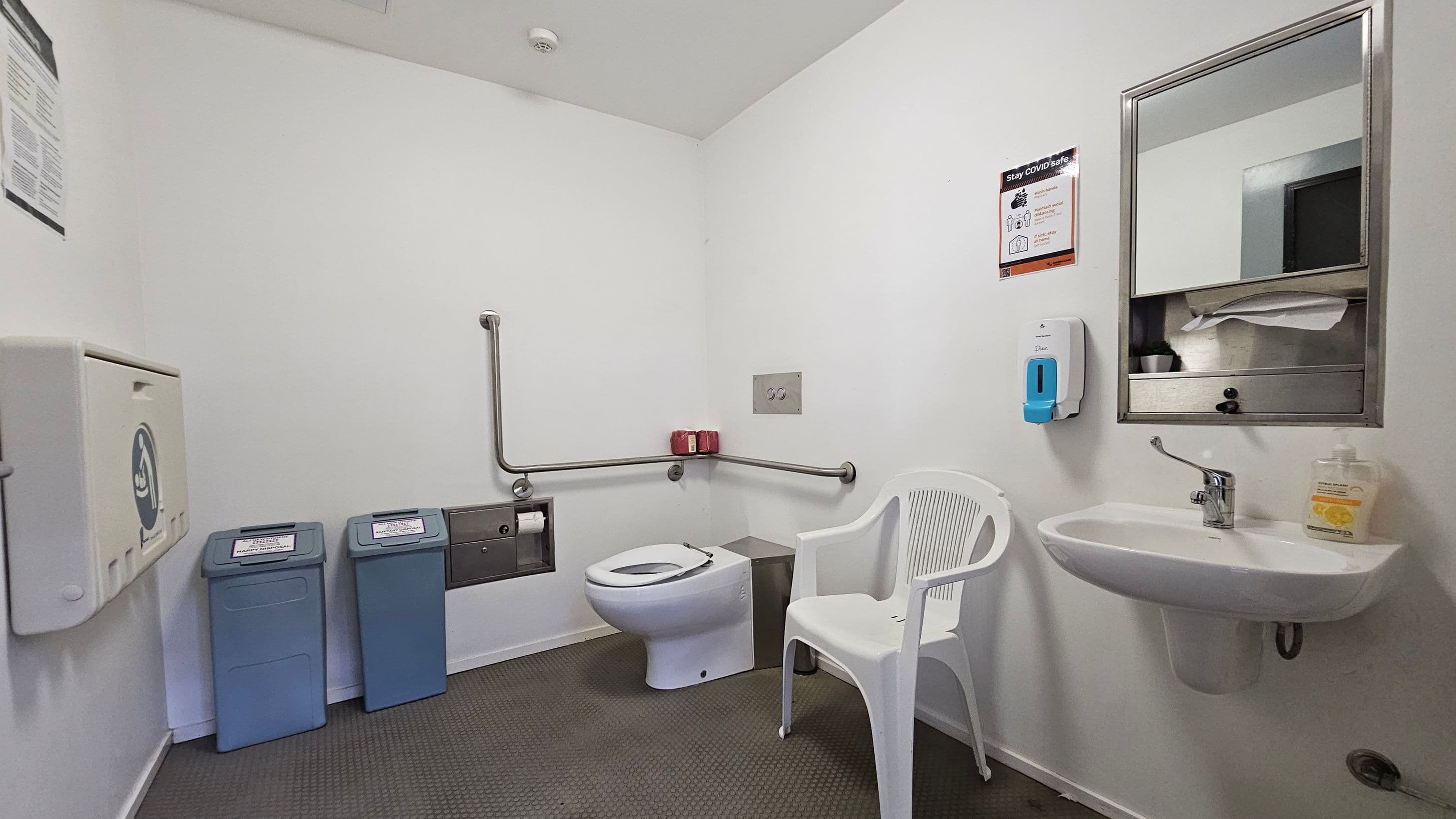 A clean, accessible bathroom with a white toilet and grab bars on the left wall. Two blue bins are positioned nearby. A white plastic chair is next to the toilet. On the right, a sink with a soap dispenser and a mirror above it. A hand sanitiser dispenser is mounted on the wall. The floor is covered with dark, textured tiles, and the walls are plain white. A "Stay COVID safe" sign is visible near the mirror.