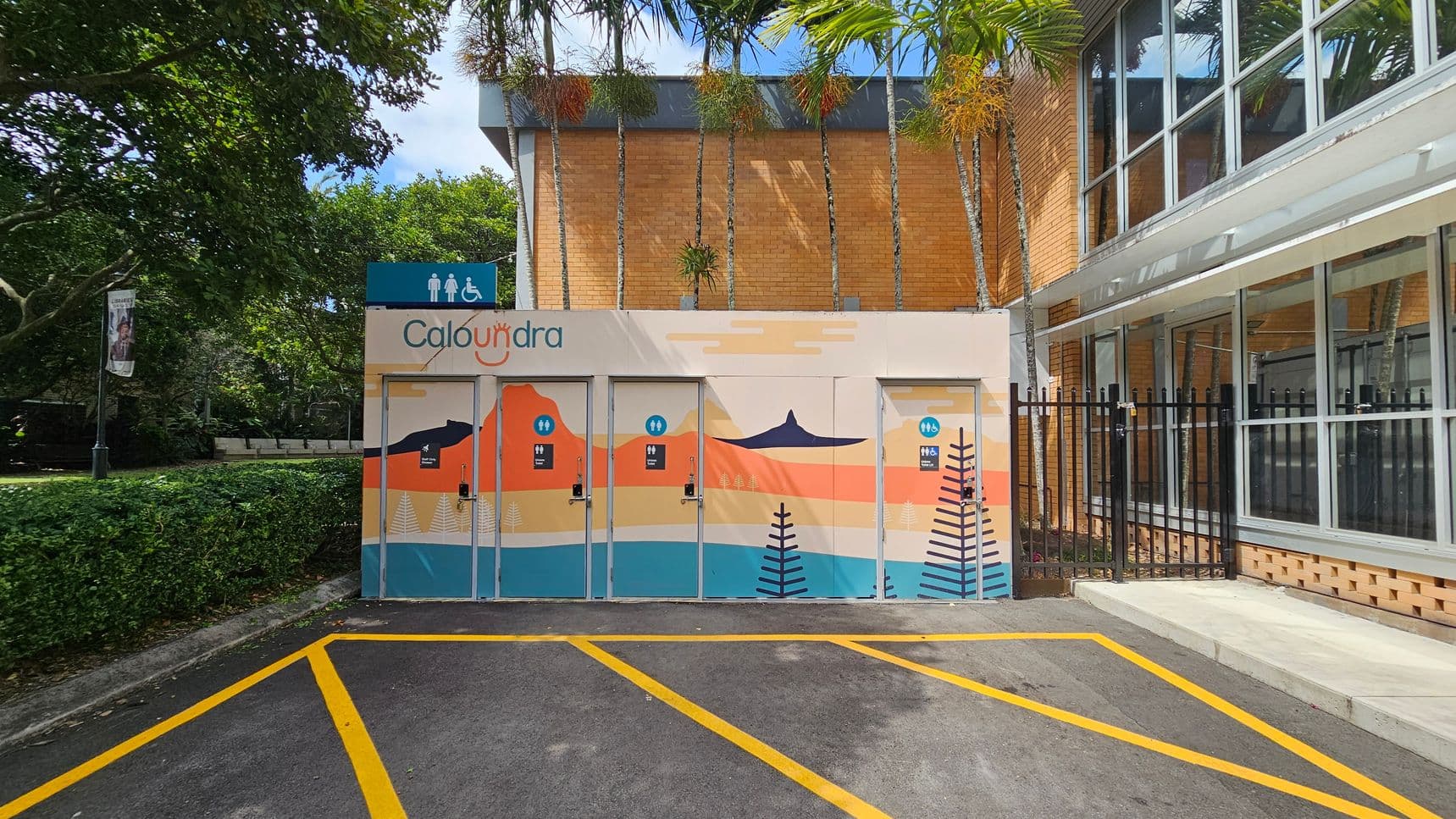 Public toilets with colourful murals are on the left, featuring abstract landscapes and the word "Caloundra." The building is made of brick with tall windows on the right. A row of palm trees is visible above the structure. In the foreground, there are parking spaces marked with yellow lines on the asphalt. A hedge and trees are on the far left, providing greenery to the scene.