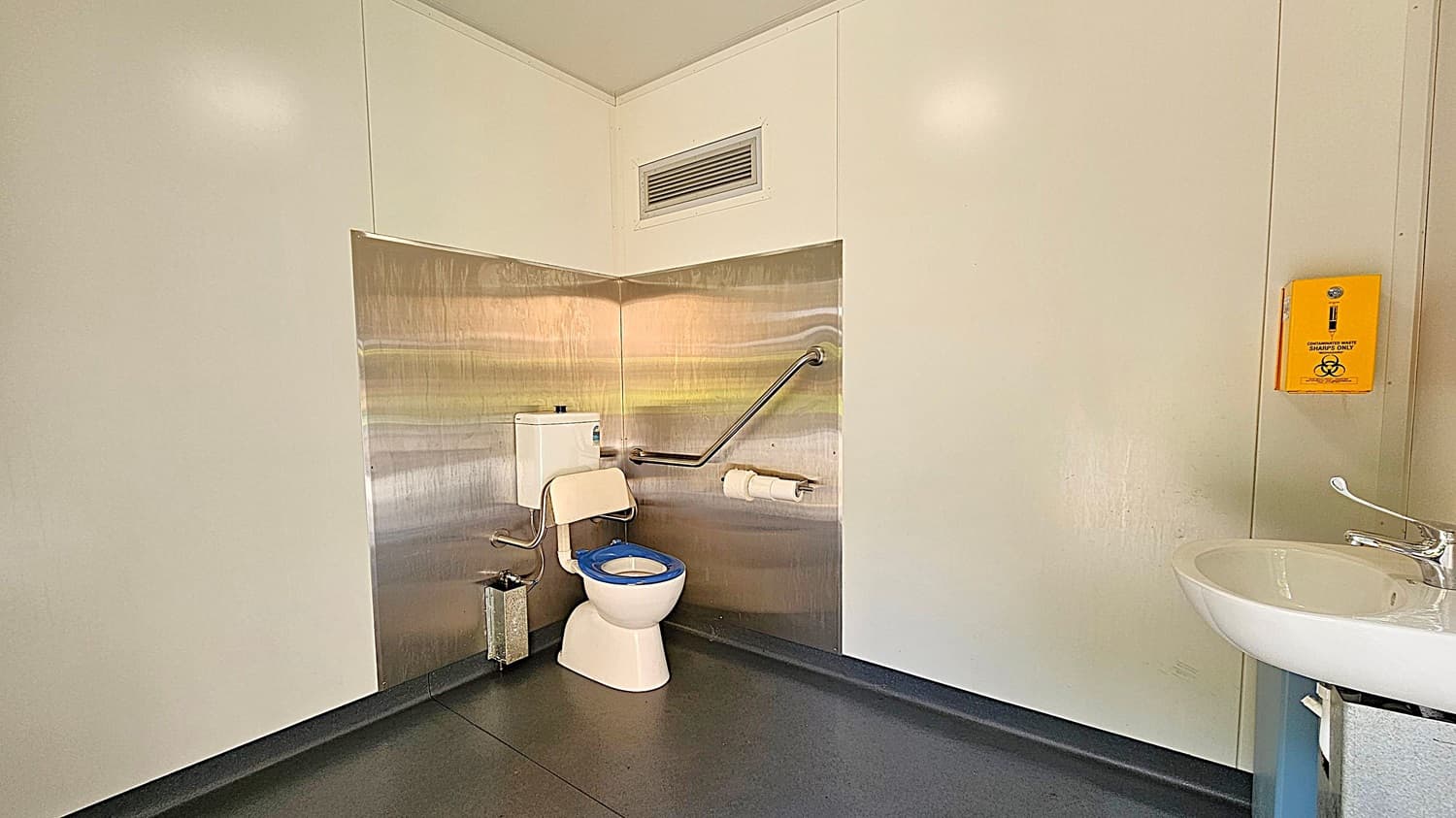 A bathroom with a white toilet featuring a blue seat is positioned against a metallic wall on the left. A grab bar and toilet paper holder are mounted on the wall. To the right, a yellow sharps disposal box is affixed to the wall. A white sink with a silver tap is partially visible in the foreground. The floor is dark grey, and the walls are light-coloured.